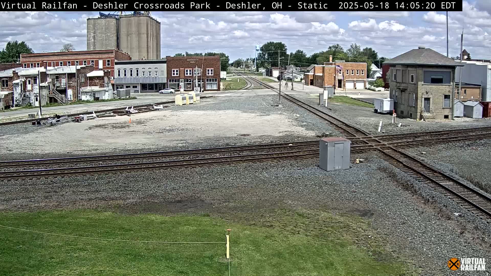 A mostly sunny day shows a railroad crossing in a small town with several buildings, grain silos, and a railway signal control tower.