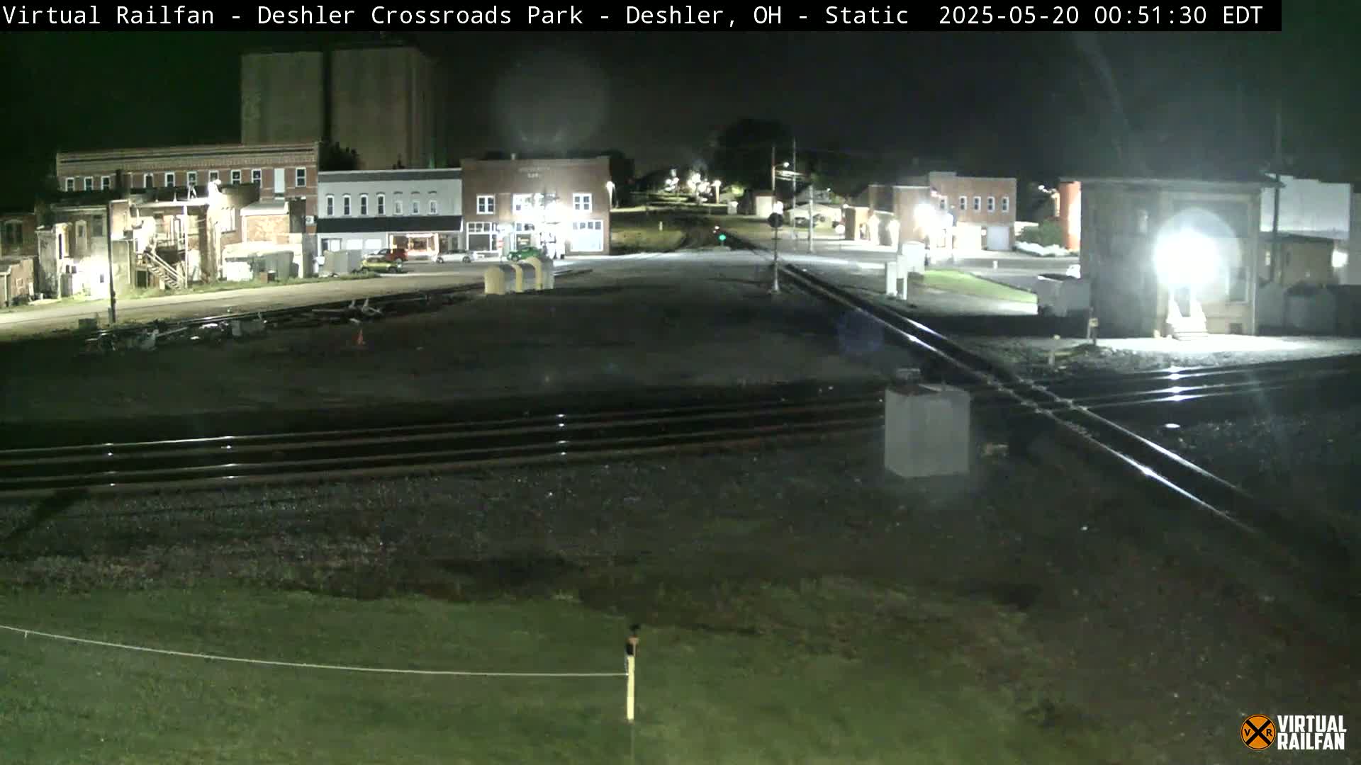 A nighttime view of a railroad crossing in a small town, with several buildings visible under dark, clear skies.