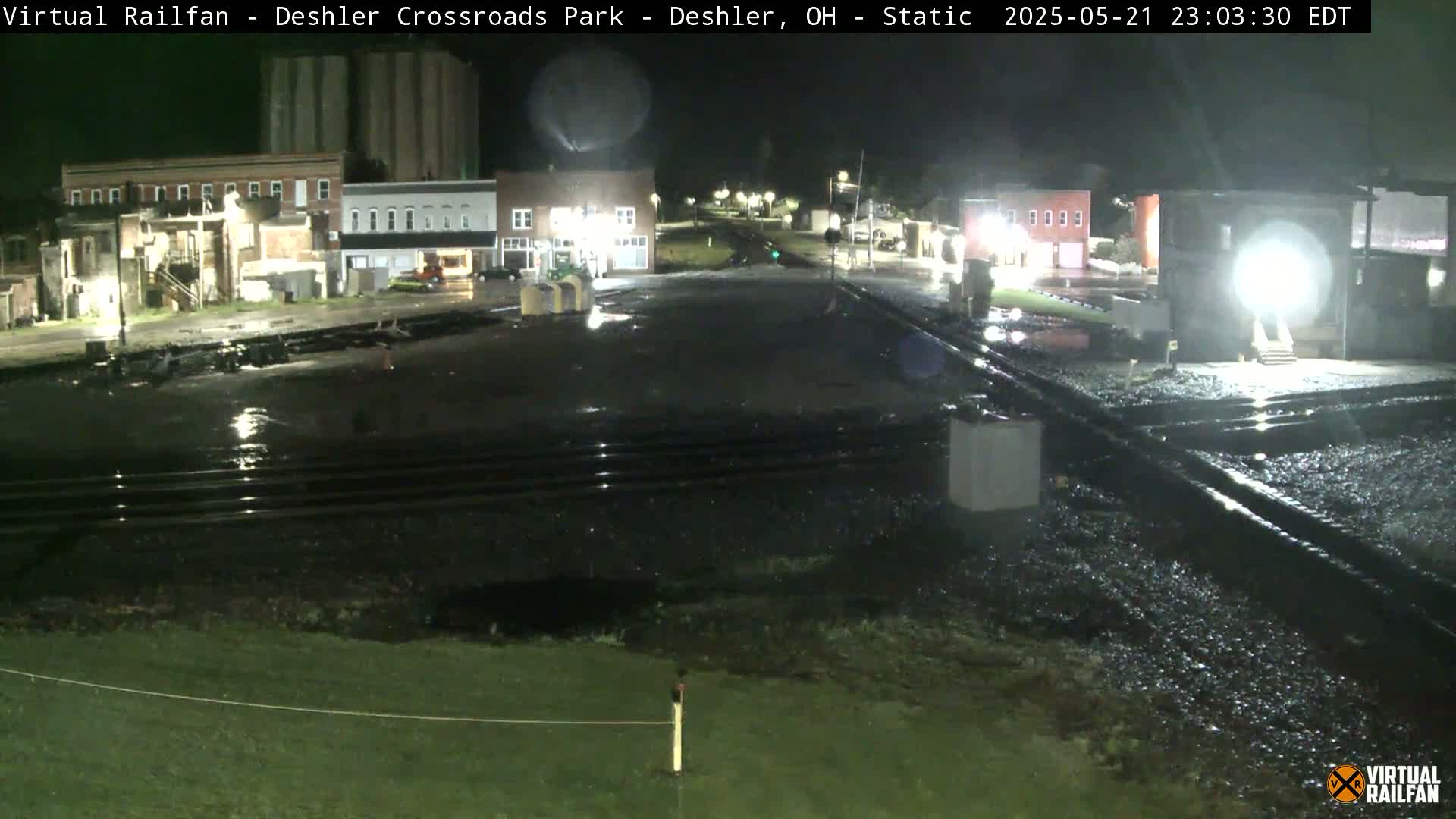 A nighttime view of a wet, empty town square with railroad tracks in the foreground, under a dark sky.