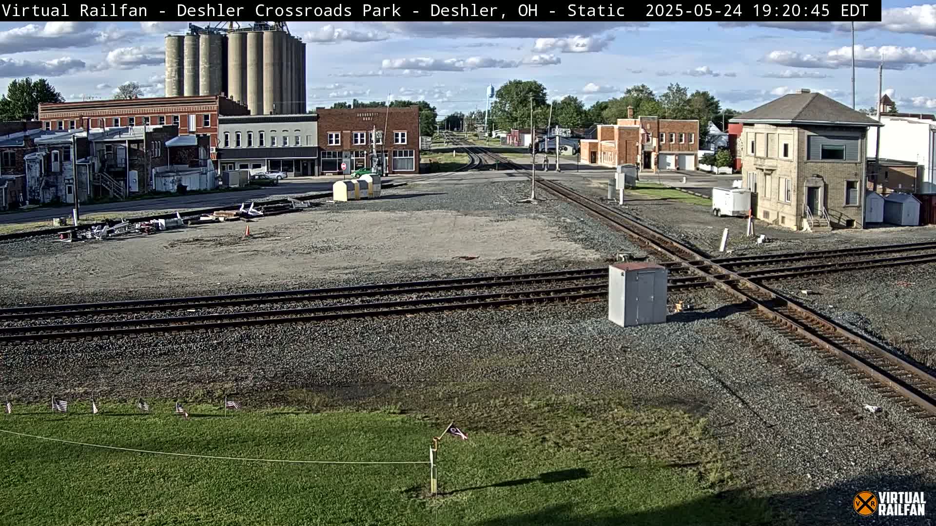 A mostly sunny view of a railroad crossing in a small town, with several buildings and grain silos visible in the background.