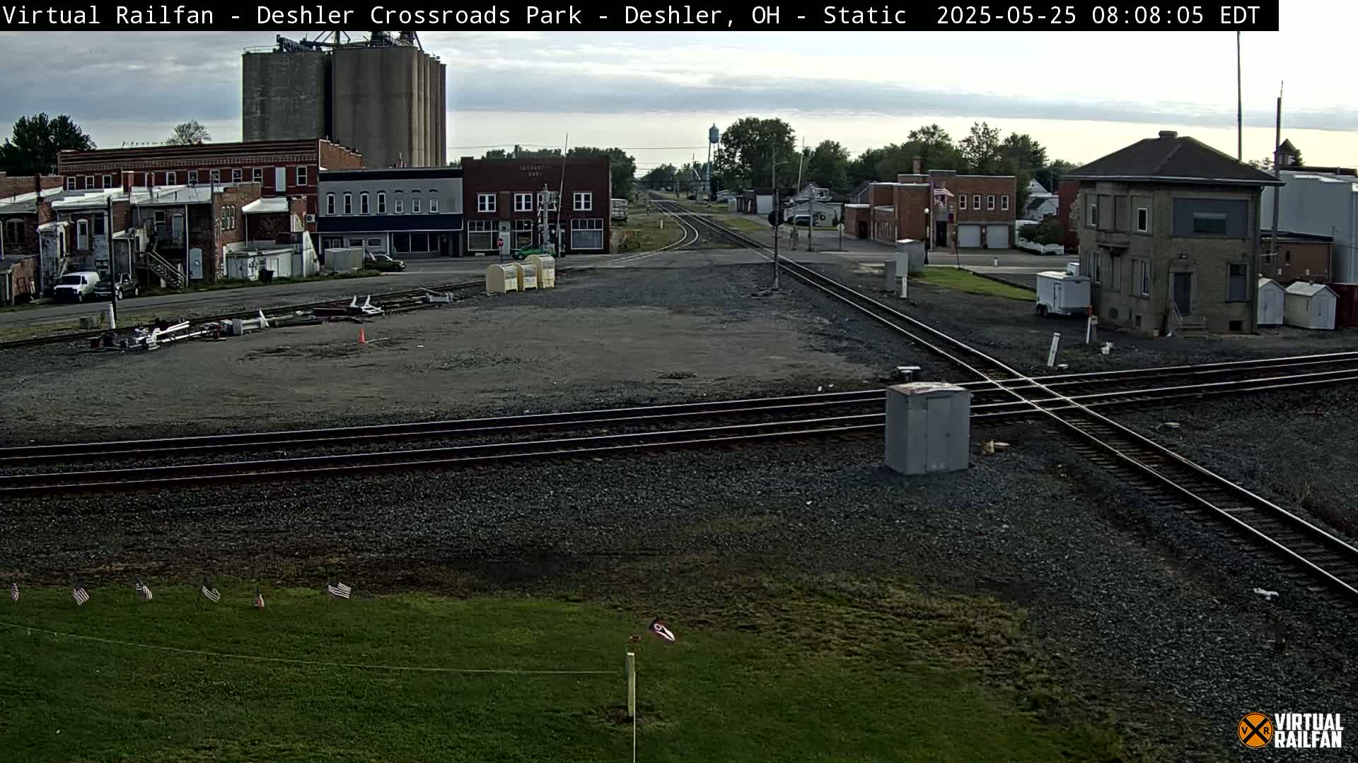 A mostly cloudy day reveals a railroad crossing in a small town with several buildings, grain silos, and a few cars.