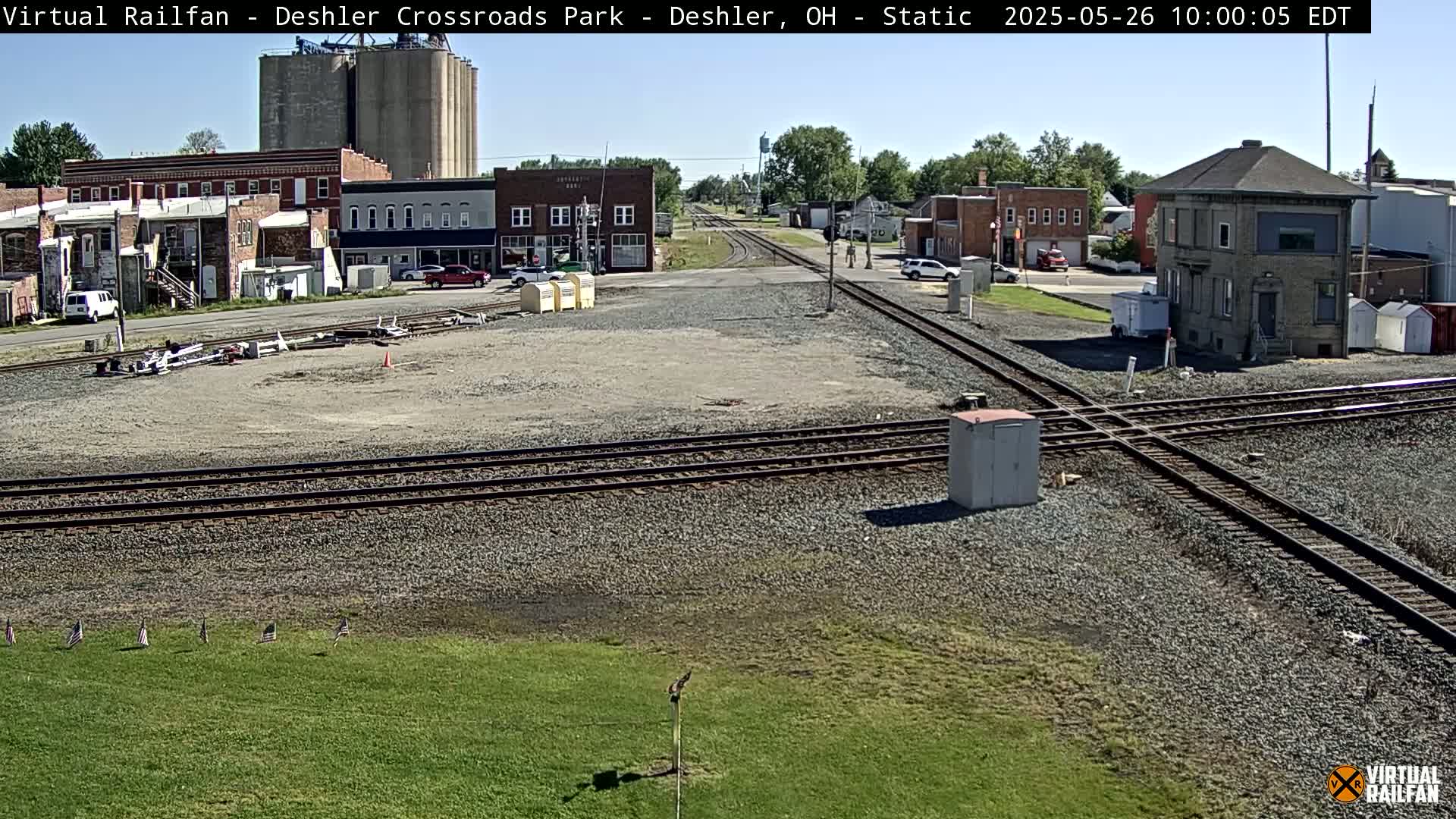 A railroad crossing in a small town on a sunny day, with several buildings and grain silos visible in the background.