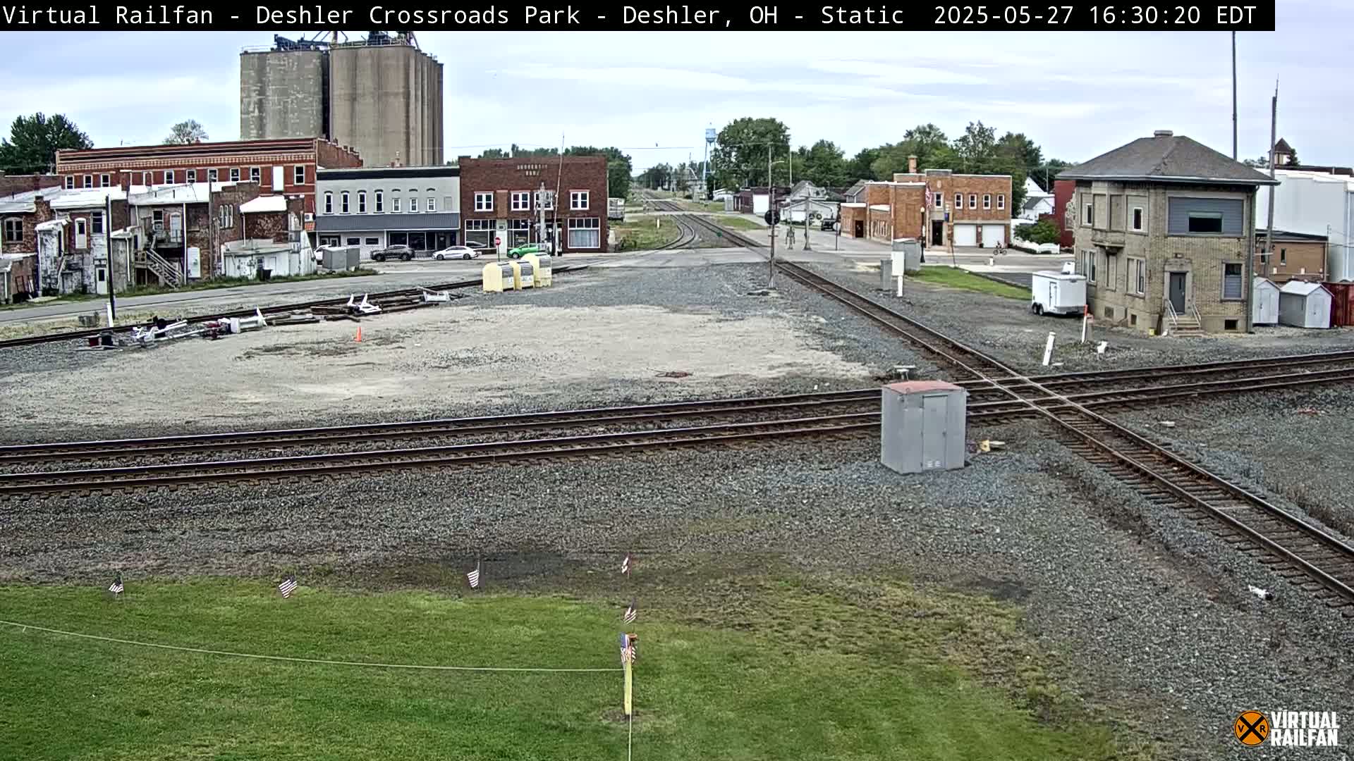 A mostly sunny view of a train crossing in a small town, with several buildings, grain silos, and a few small American flags in the foreground.