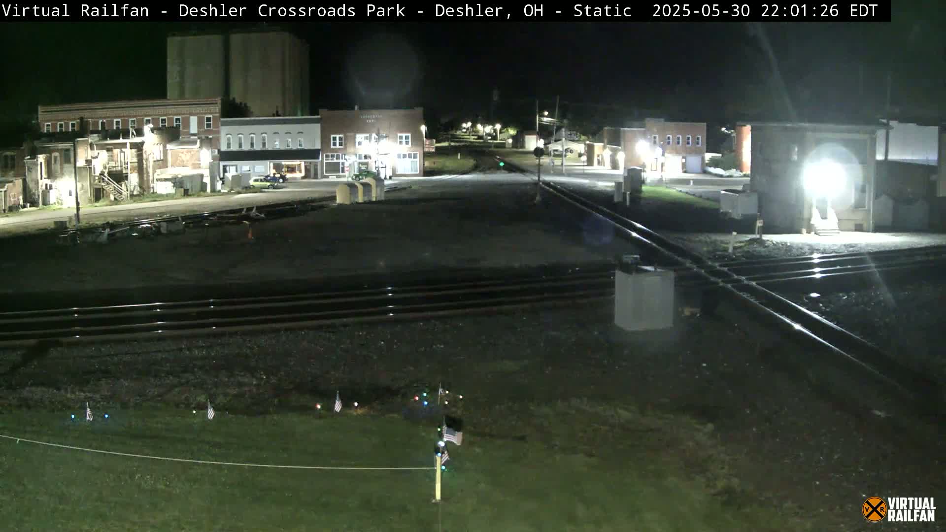 A nighttime view of a railroad crossing in a small town, with several buildings visible in the background.