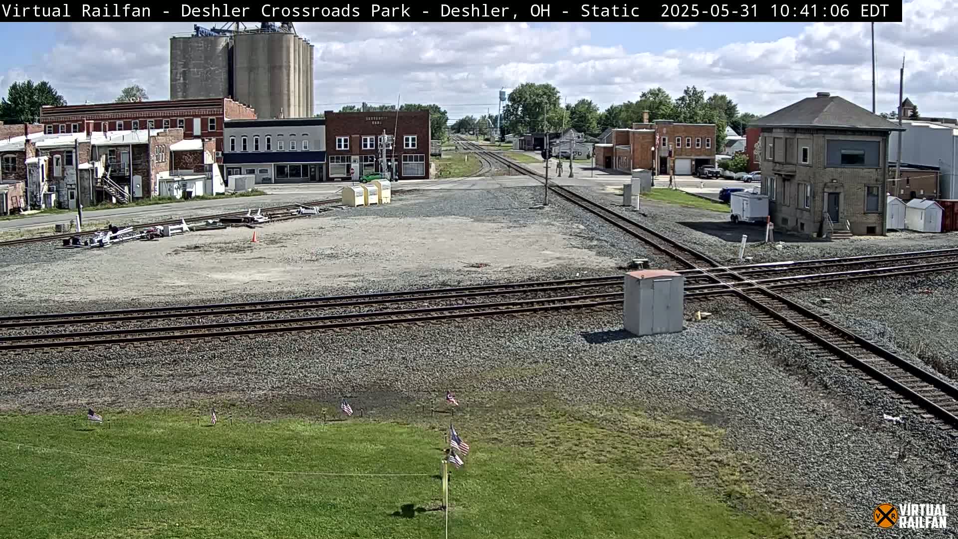 A mostly sunny day shows a railroad crossing in a small town, with several buildings, grain silos, and a few American flags in the foreground.