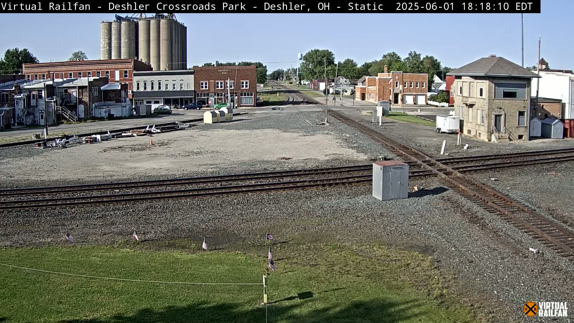 A sunny view of a railroad crossing in a small town, with several grain silos and buildings visible in the background.