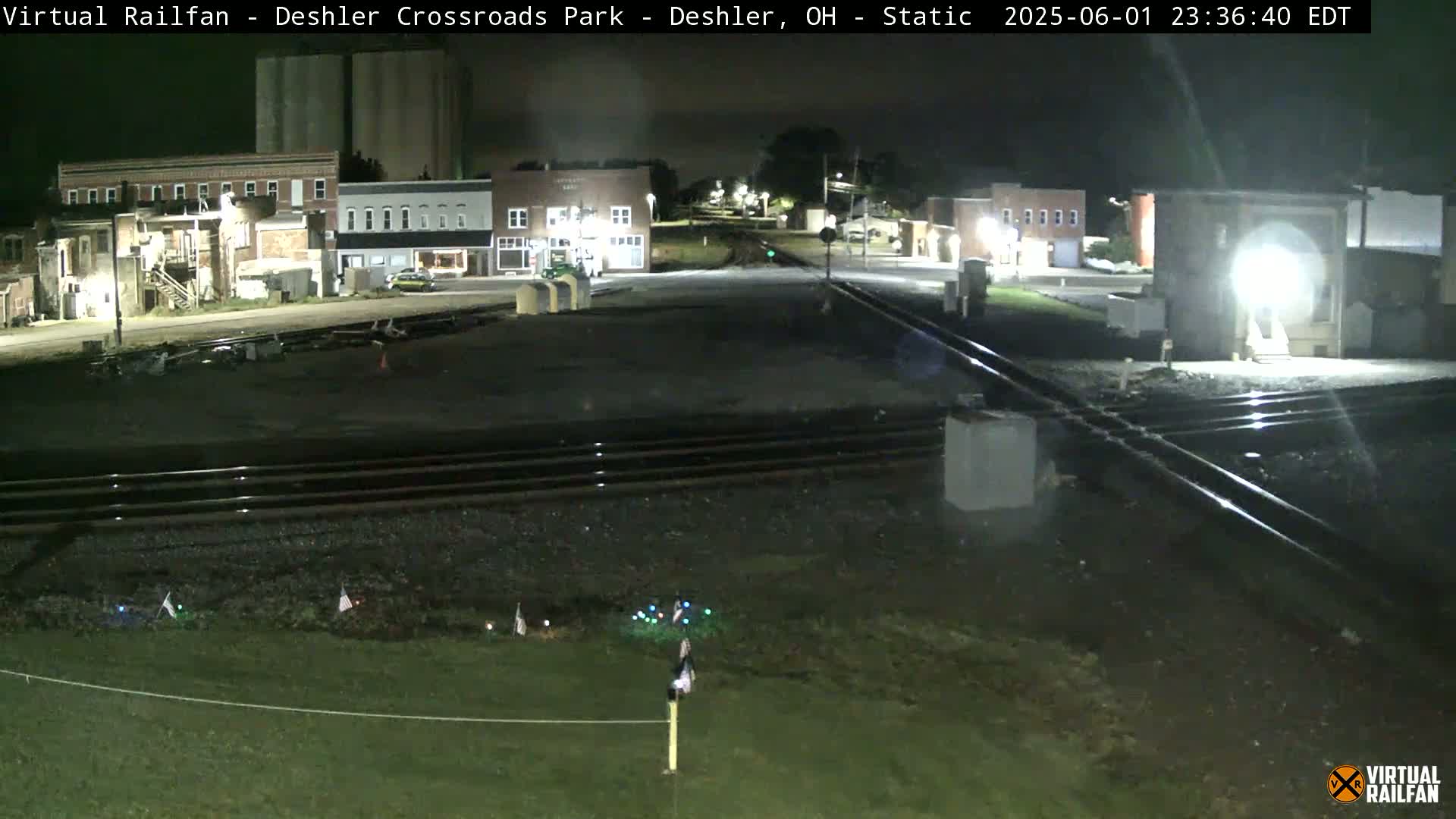A nighttime view of a railroad crossing near several brick buildings, with some small flags and lights visible in the foreground.