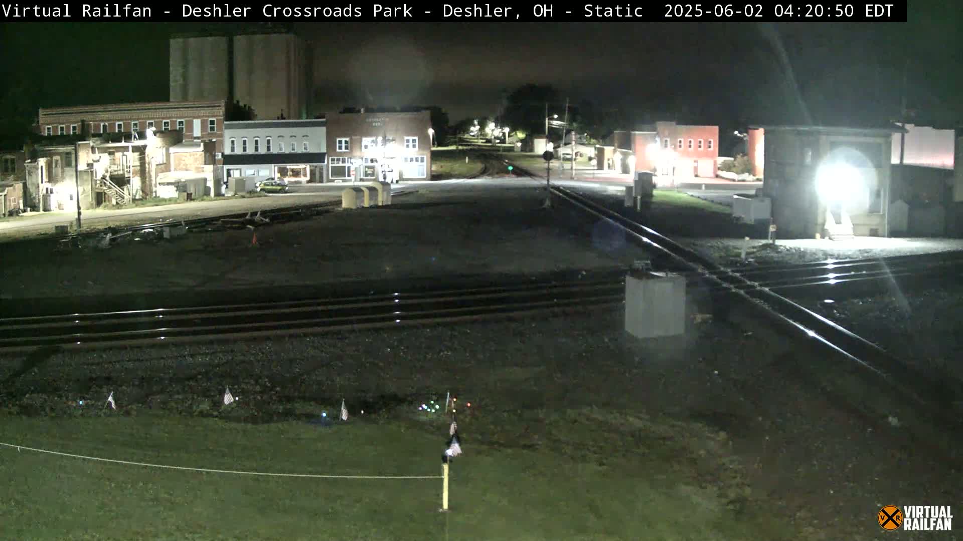 A nighttime view of a railroad crossing in a small town, with several brick buildings visible in the background under dark skies.