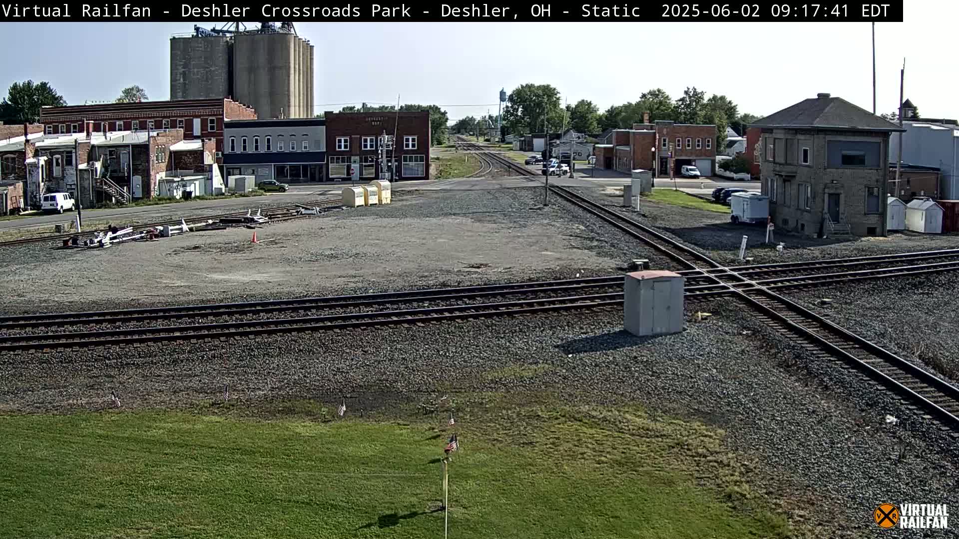 A sunny view of a railroad crossing in a small town, with several buildings and grain silos in the background.