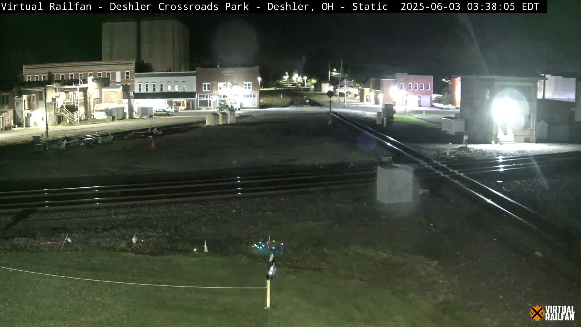 A nighttime view of a railroad crossing near several buildings, with the ground appearing damp.