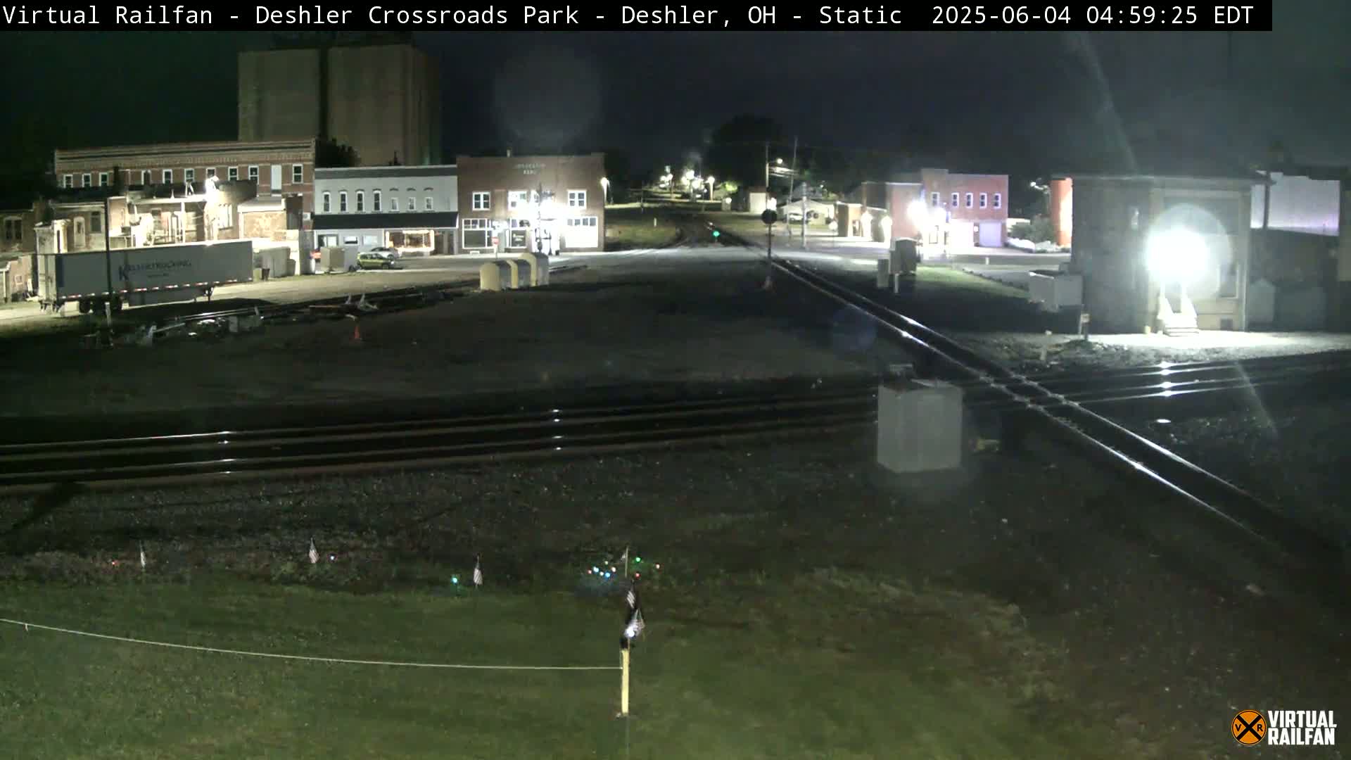 A nighttime view of a railroad crossing in a small town, with several buildings visible under artificial light.