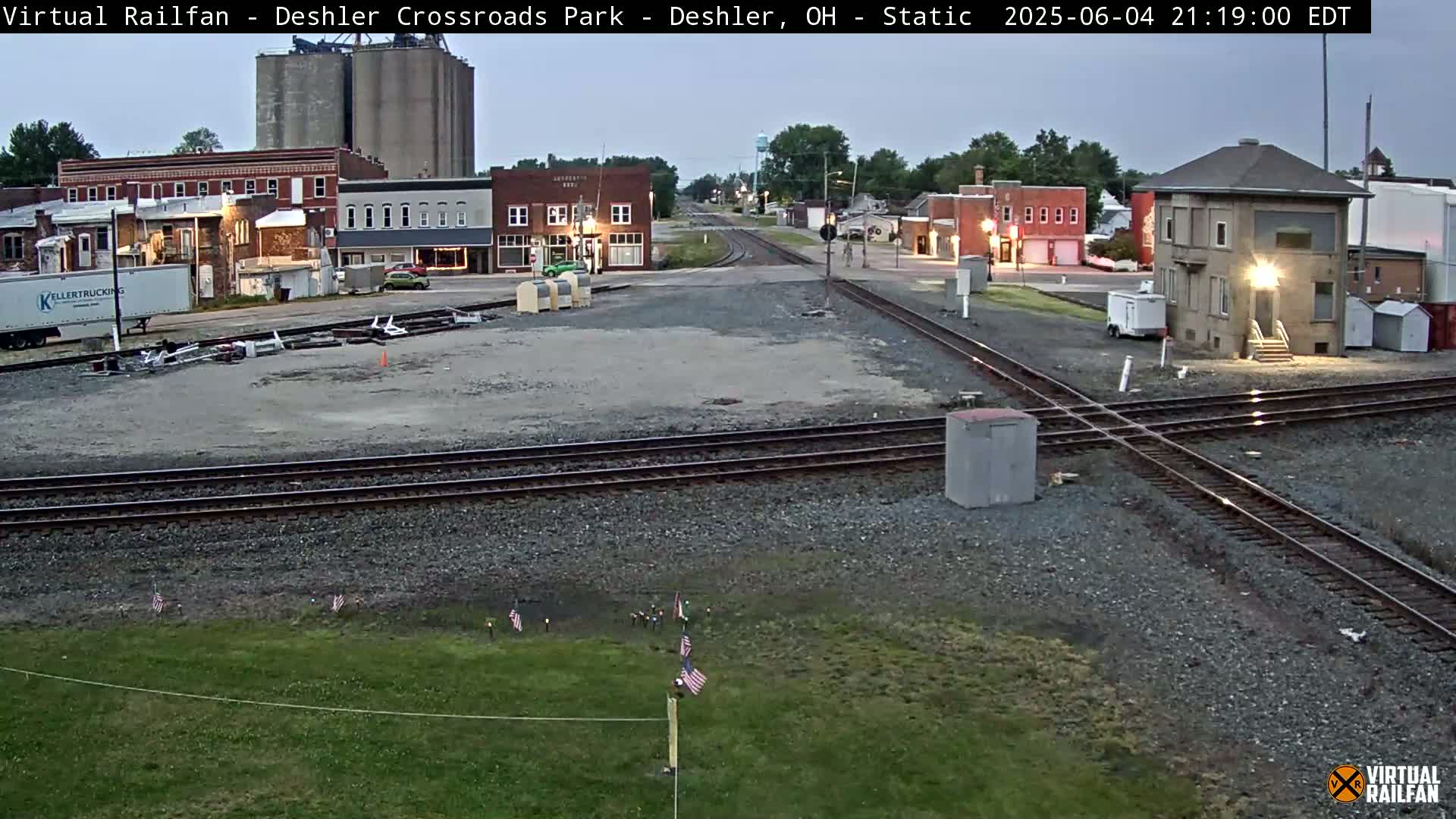 A nighttime view of a railroad crossing in a small town, with several buildings and grain silos visible under a twilight sky.