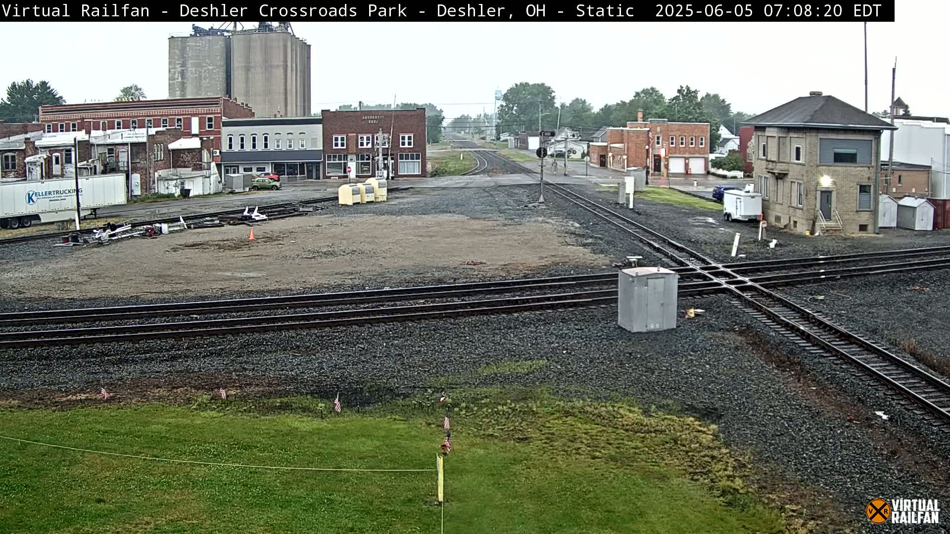 A railroad crossing in a small town on an overcast day, with several buildings, including a grain silo and a small brick structure, visible in the background.