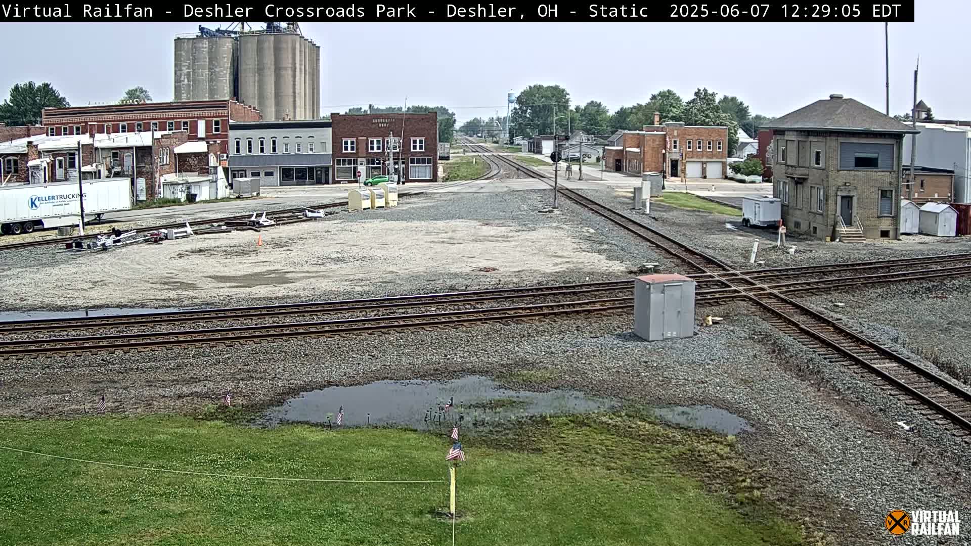 A railroad crossing in a small town on an overcast day, with several buildings visible in the background and puddles of water near the tracks.