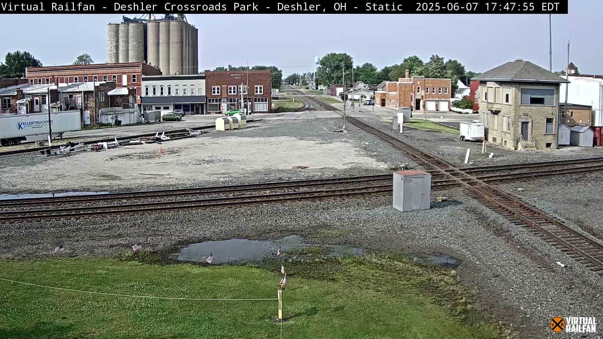A sunny view of a railroad crossing in a small town, showing several buildings, grain silos, and a train signal tower.