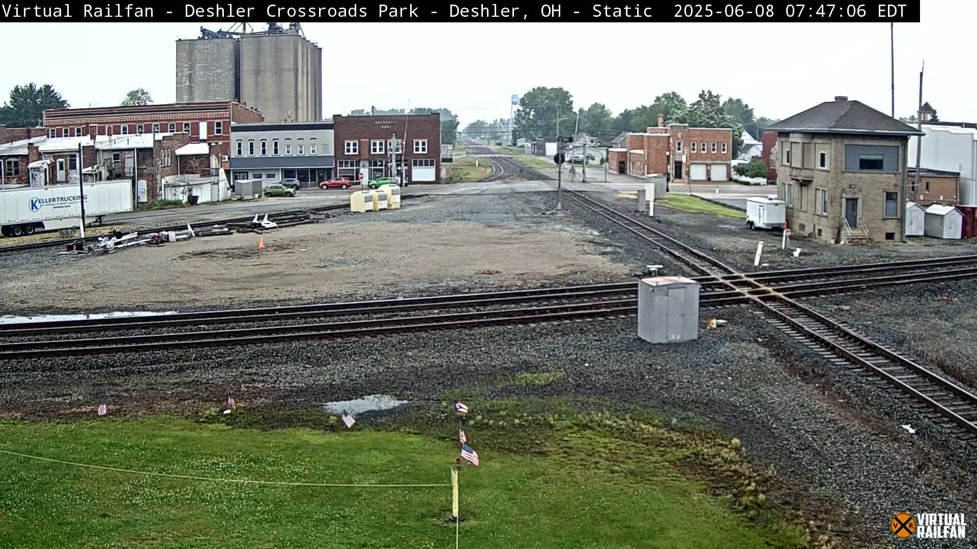 A railroad crossing in a small town on an overcast day, with several buildings, grain silos, and a small number of cars visible in the background.