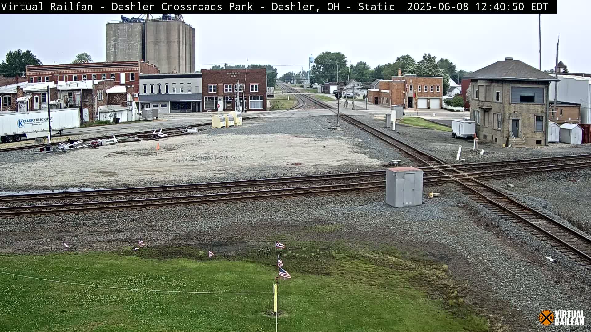 A railway crossing in a small town on an overcast day, with several buildings, grain silos, and a large semi-trailer truck visible.