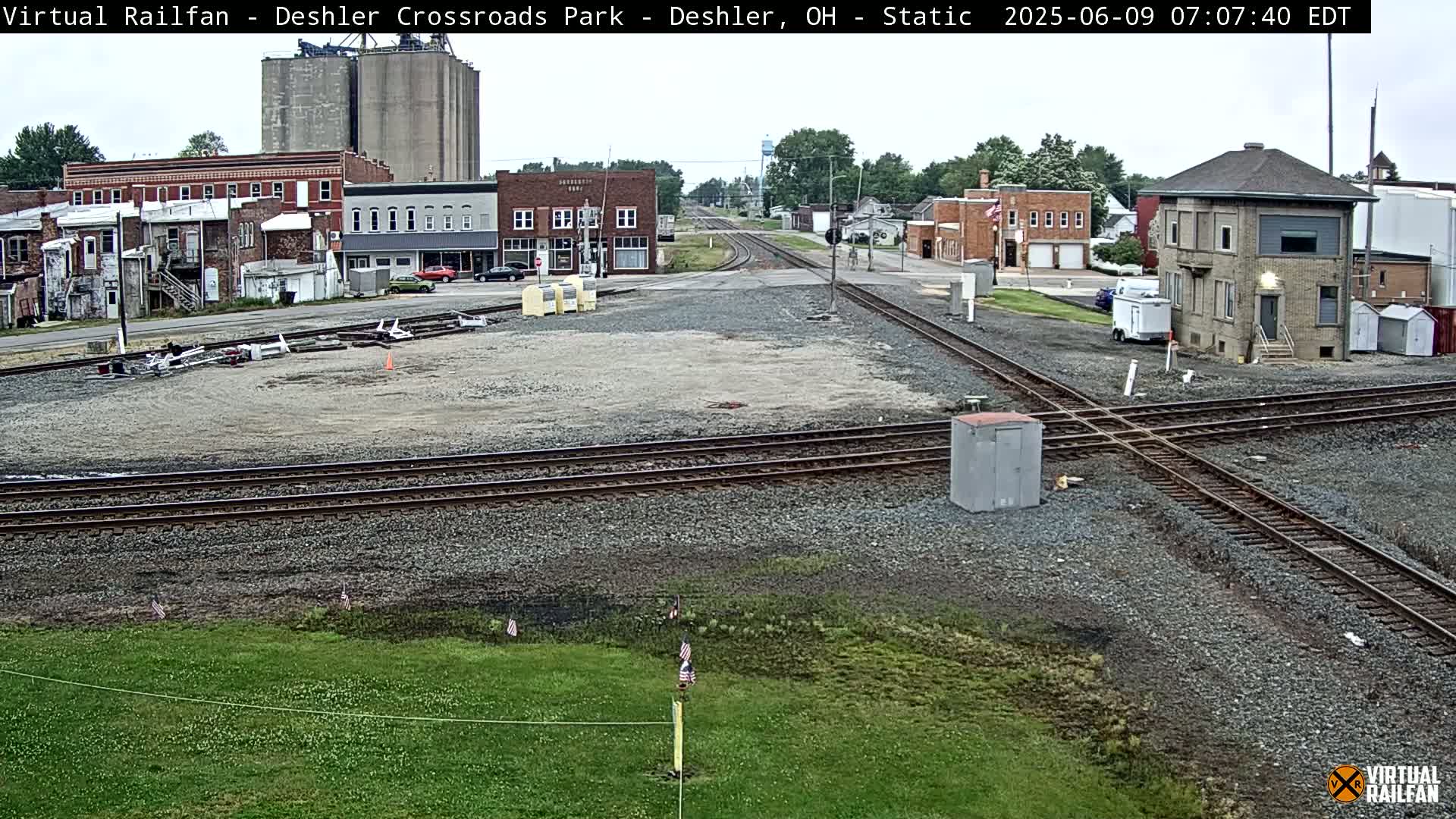 A railroad crossing in a small town on an overcast day, with several buildings and grain silos in the background.