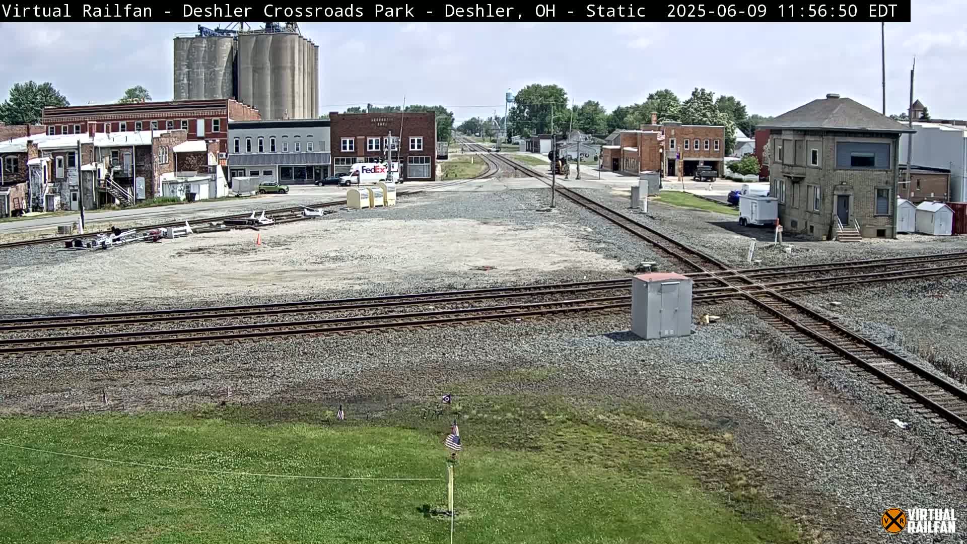 A railroad crossing in a small town on a partly sunny day, with several buildings, grain silos, and a control tower visible.