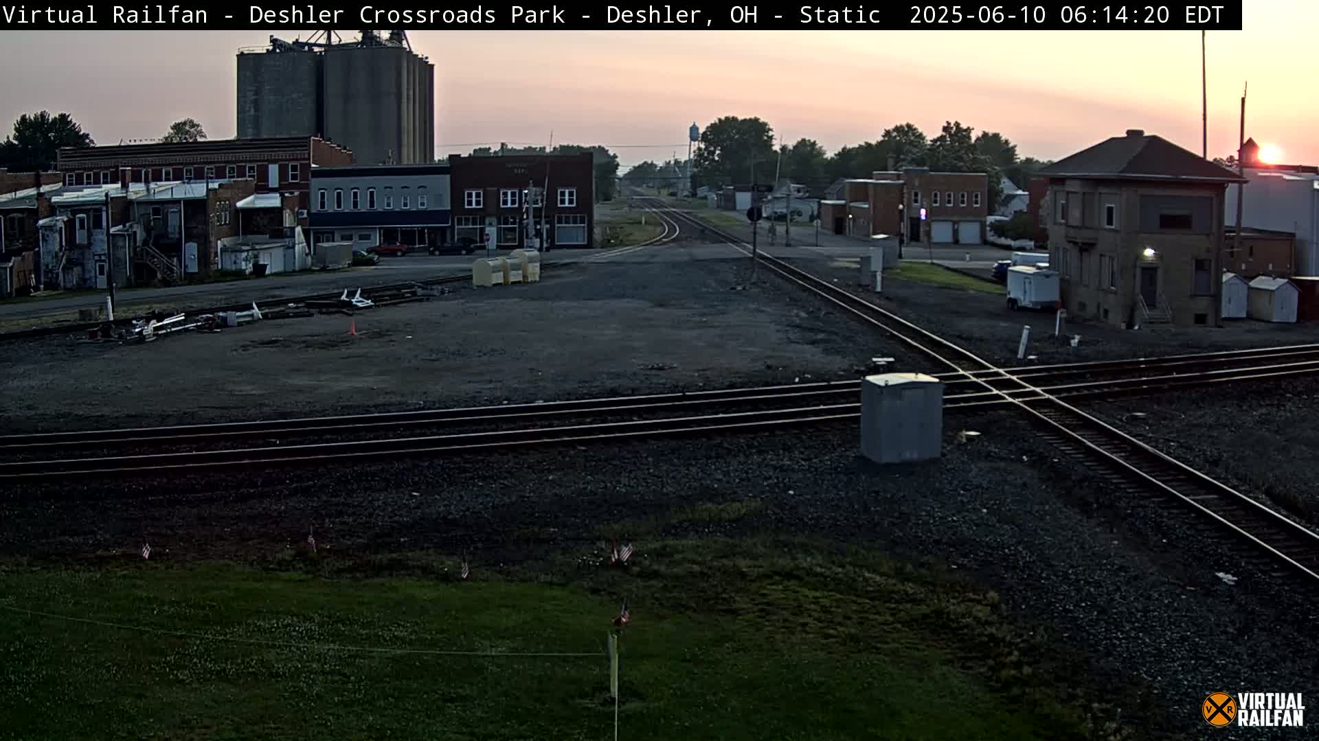 A sunrise illuminates a railroad crossing in a small town, with several buildings and grain silos visible in the background.