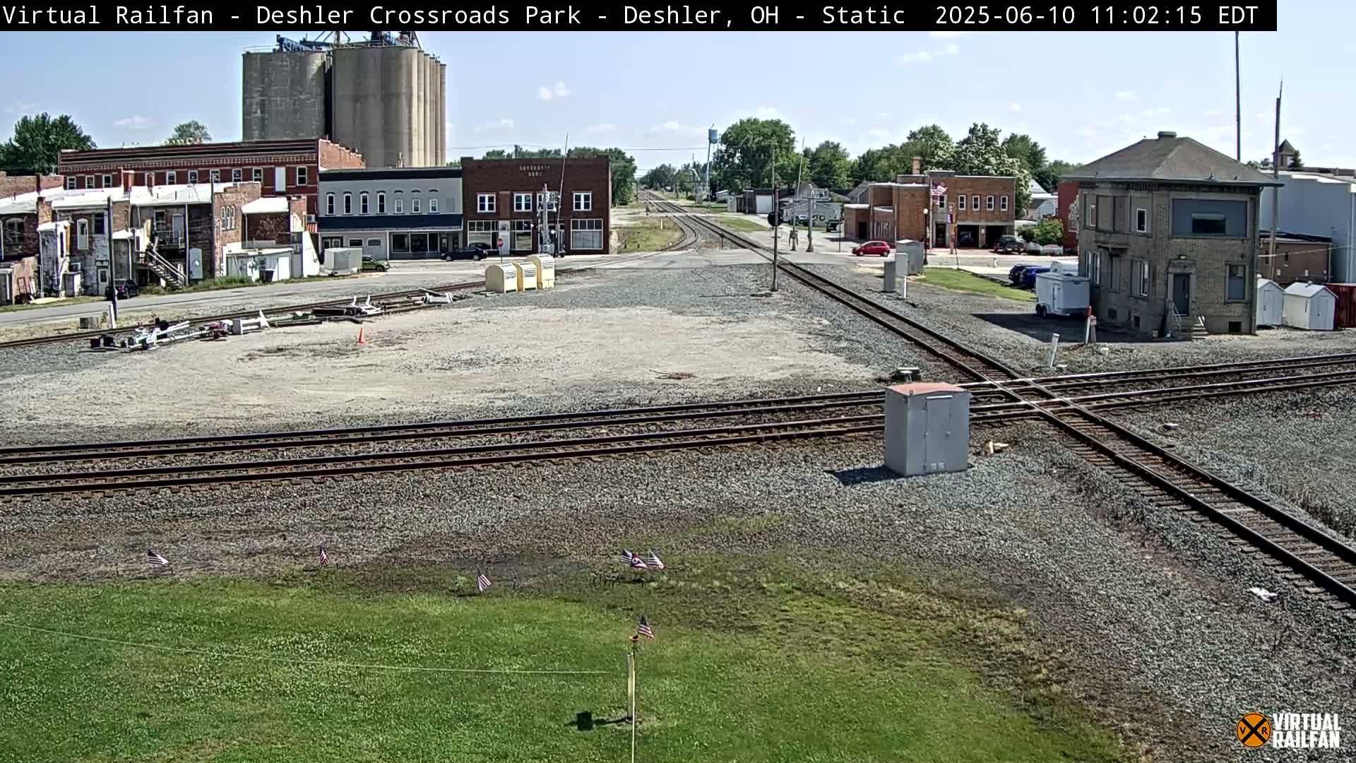 A sunny view of a railroad crossing in a small town, with several buildings, grain silos, and some cars visible.
