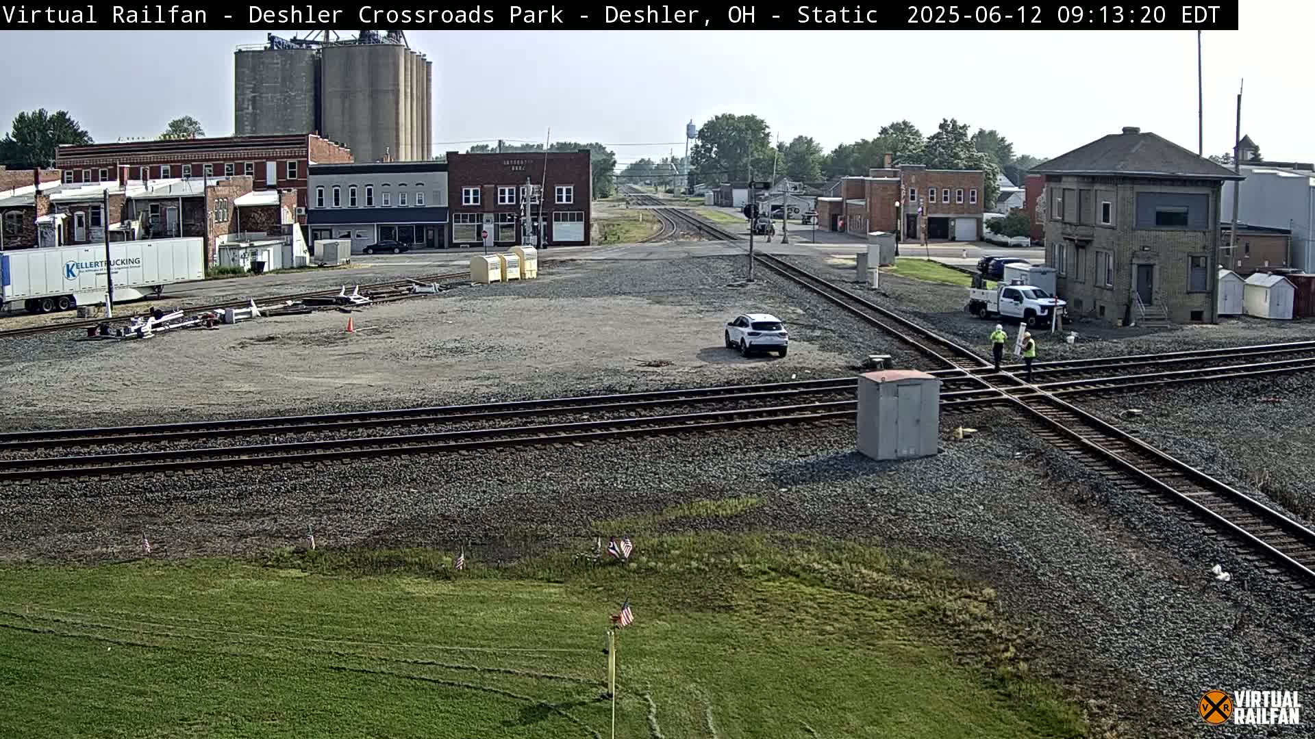 A mostly sunny view of a train crossing in a small town, with a few cars and two workers visible near the tracks.