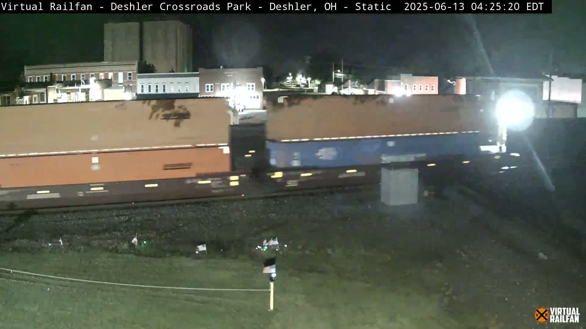 A long freight train passes by at night, near some buildings under a dark sky.