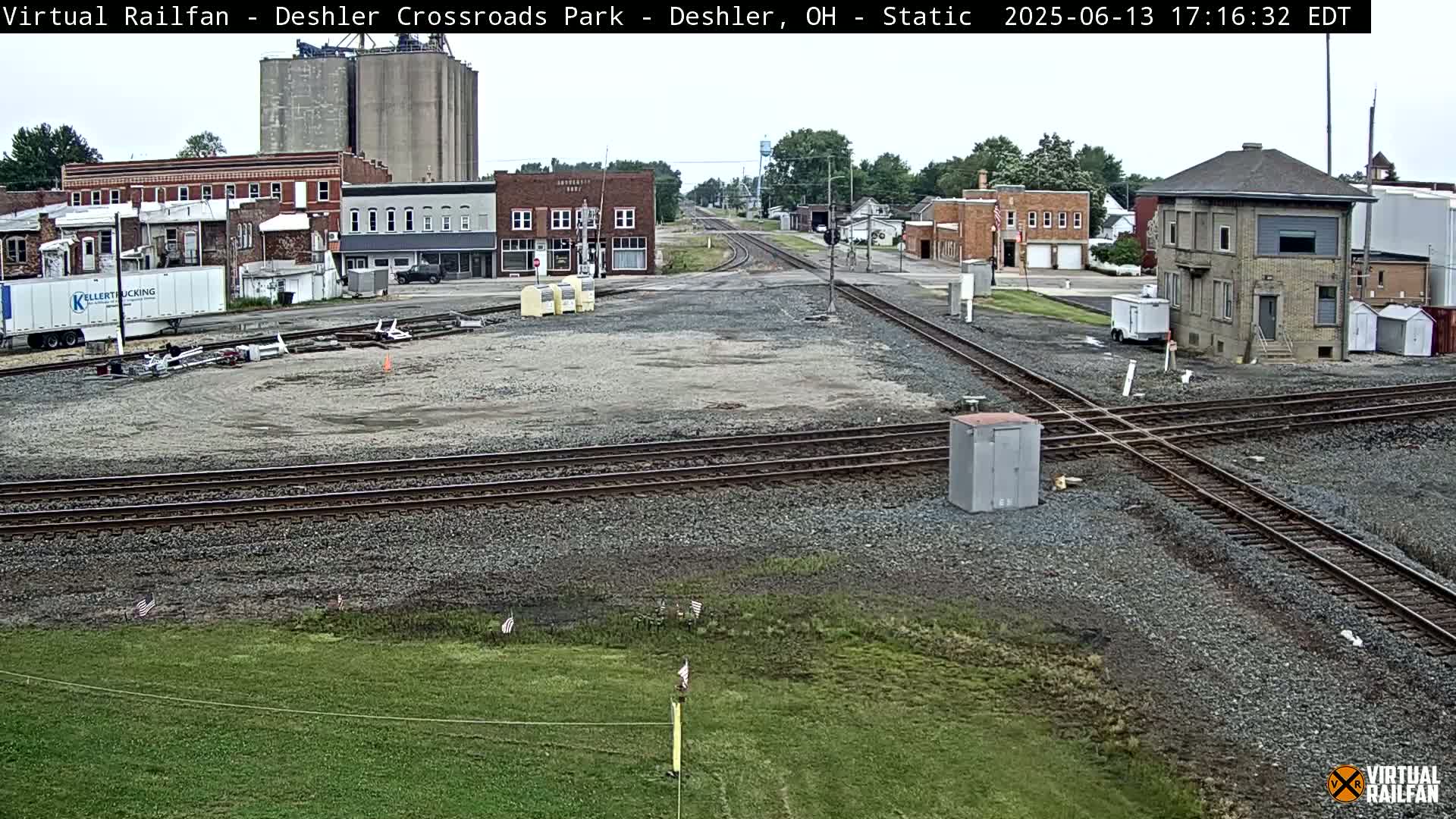 A railroad crossing in a small town on an overcast day, with buildings and grain silos in the background.
