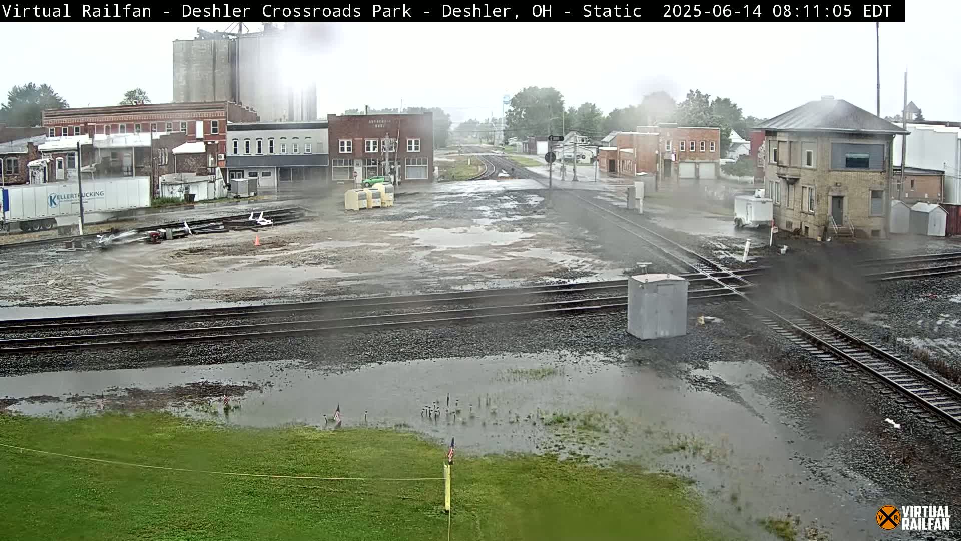A rain-soaked railroad crossing in a small town, with puddles surrounding the tracks and buildings visible in the background.