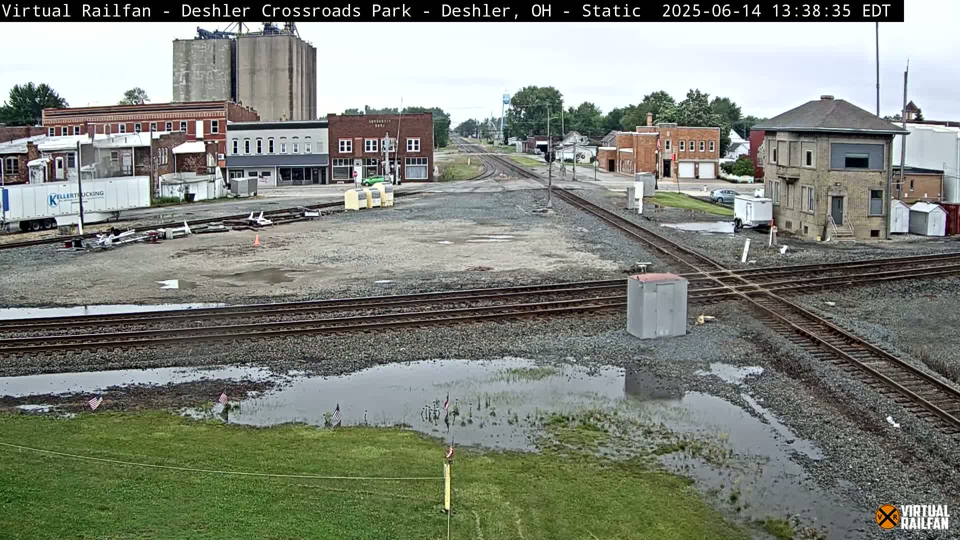 A train crossing in a small town shows several puddles of water on the ground after a light rain, with buildings and grain silos in the background.
