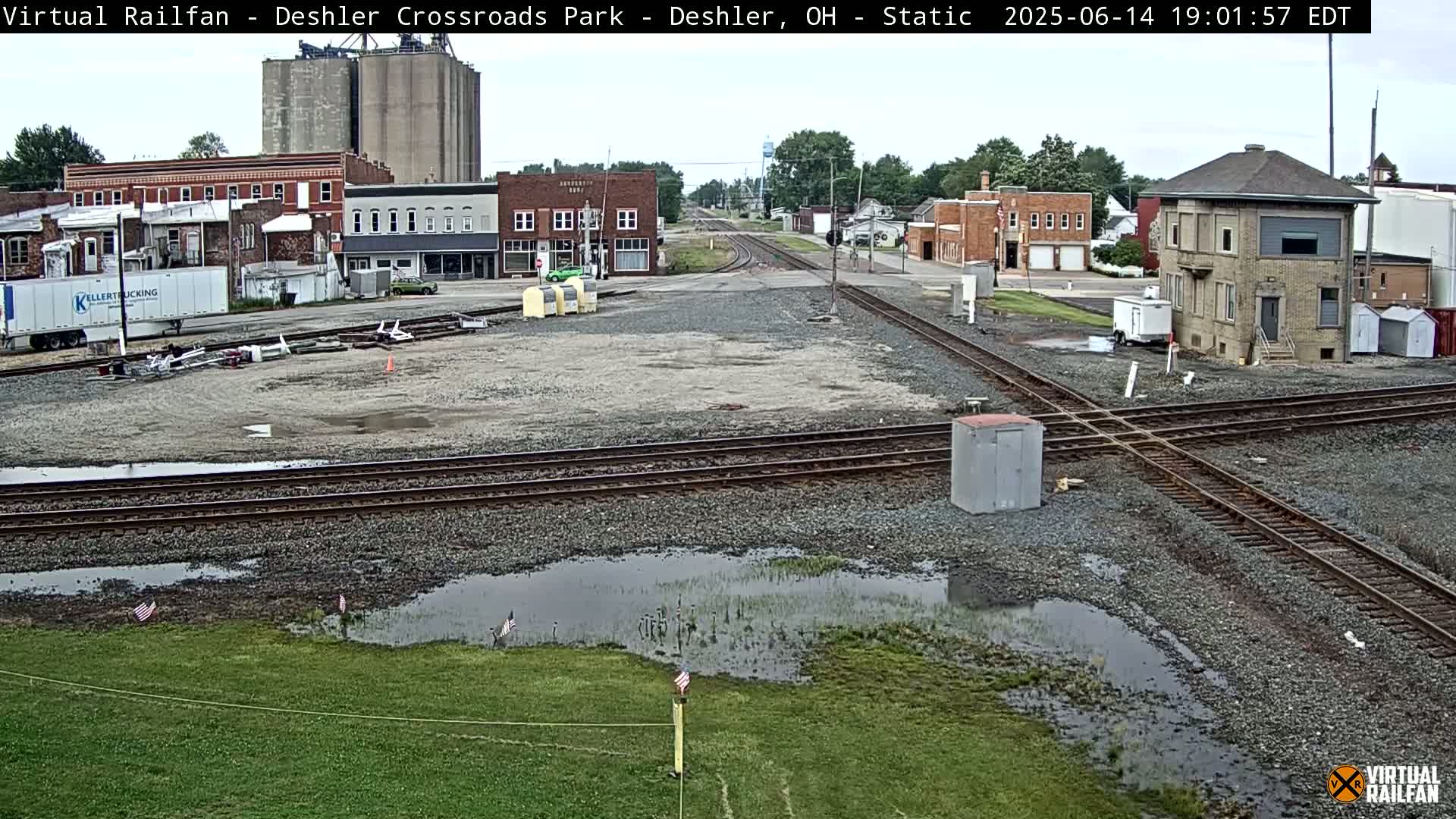 A train crossing in a small town shows several puddles of water on the ground after a light rain, with buildings and grain silos in the background.