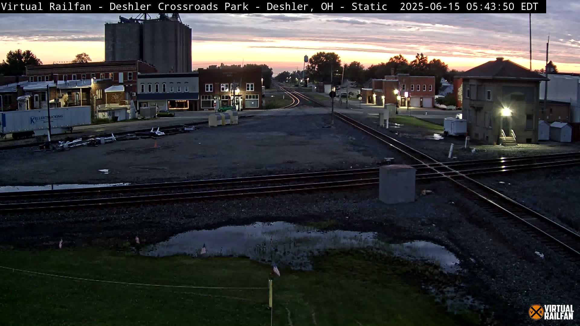 A sunrise view of a railway crossing in a small town, showing buildings, tracks, and puddles of water.