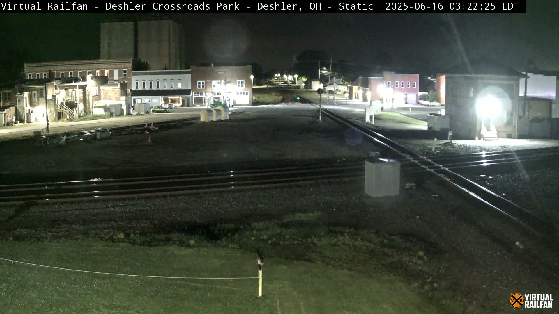 At night, a train crossing in a small town is shown with several buildings visible in the background.