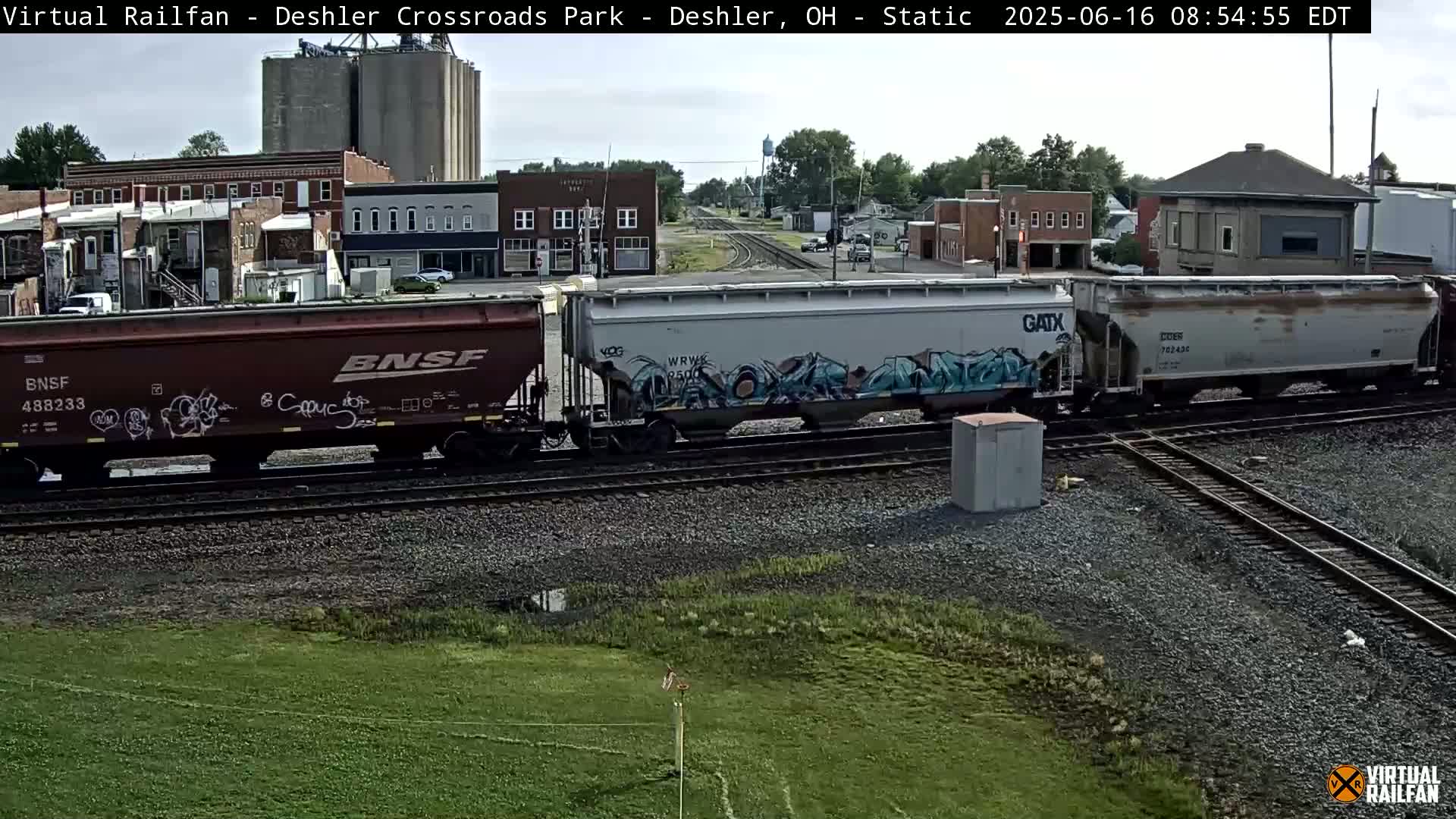 A train with graffiti-covered freight cars sits on railway tracks in front of a small town under a partly cloudy sky.
