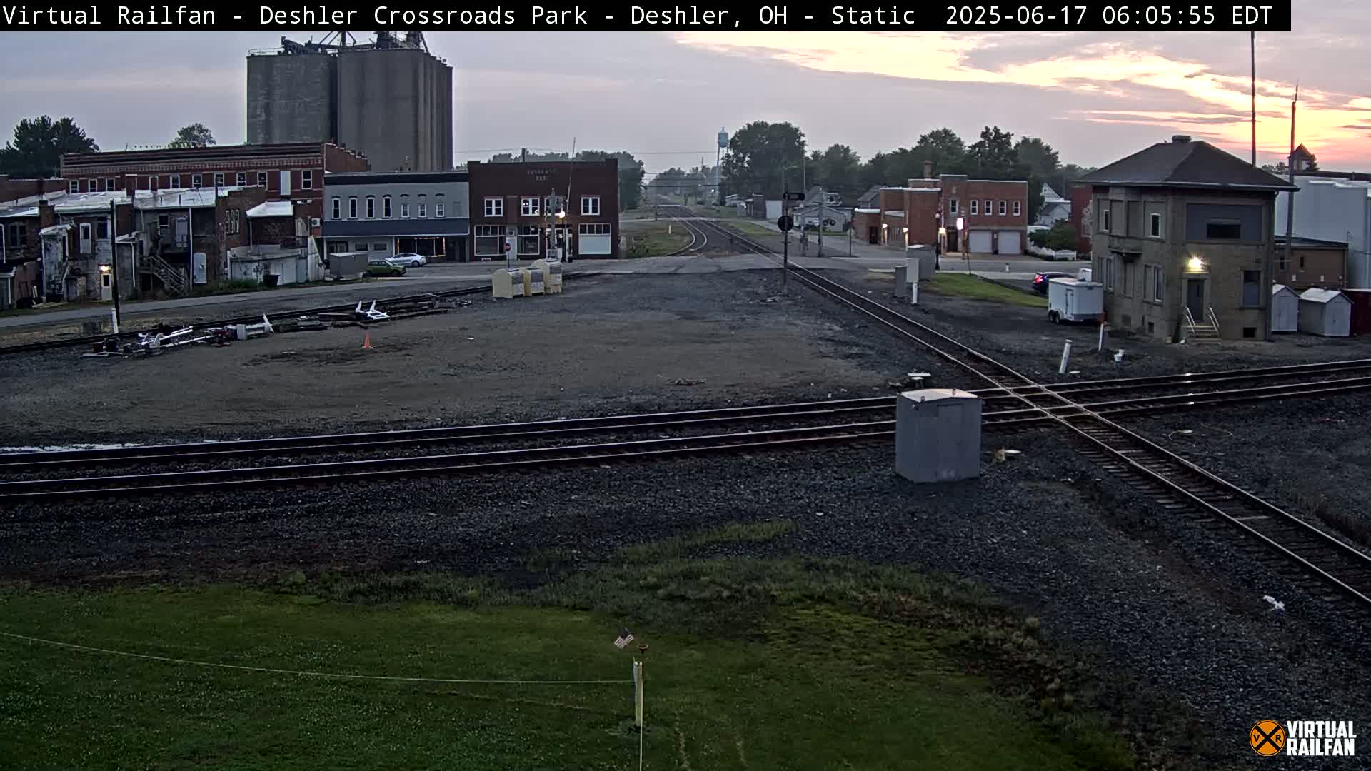 A railroad crossing in a small town at dawn, with several buildings and grain silos visible in the background.