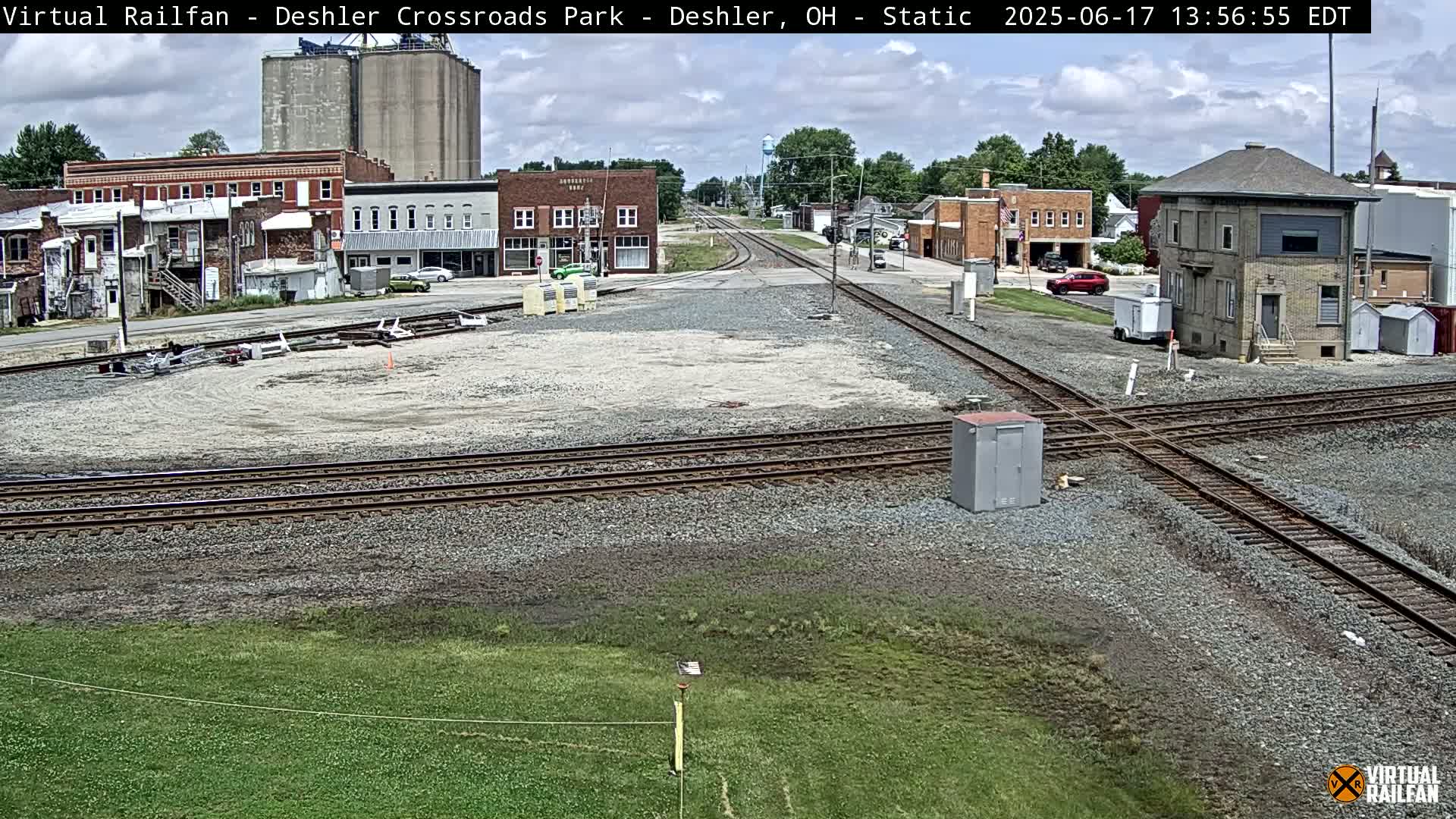 A mostly sunny view of a railroad crossing in a small town, featuring several buildings, train tracks, and a small grassy area.