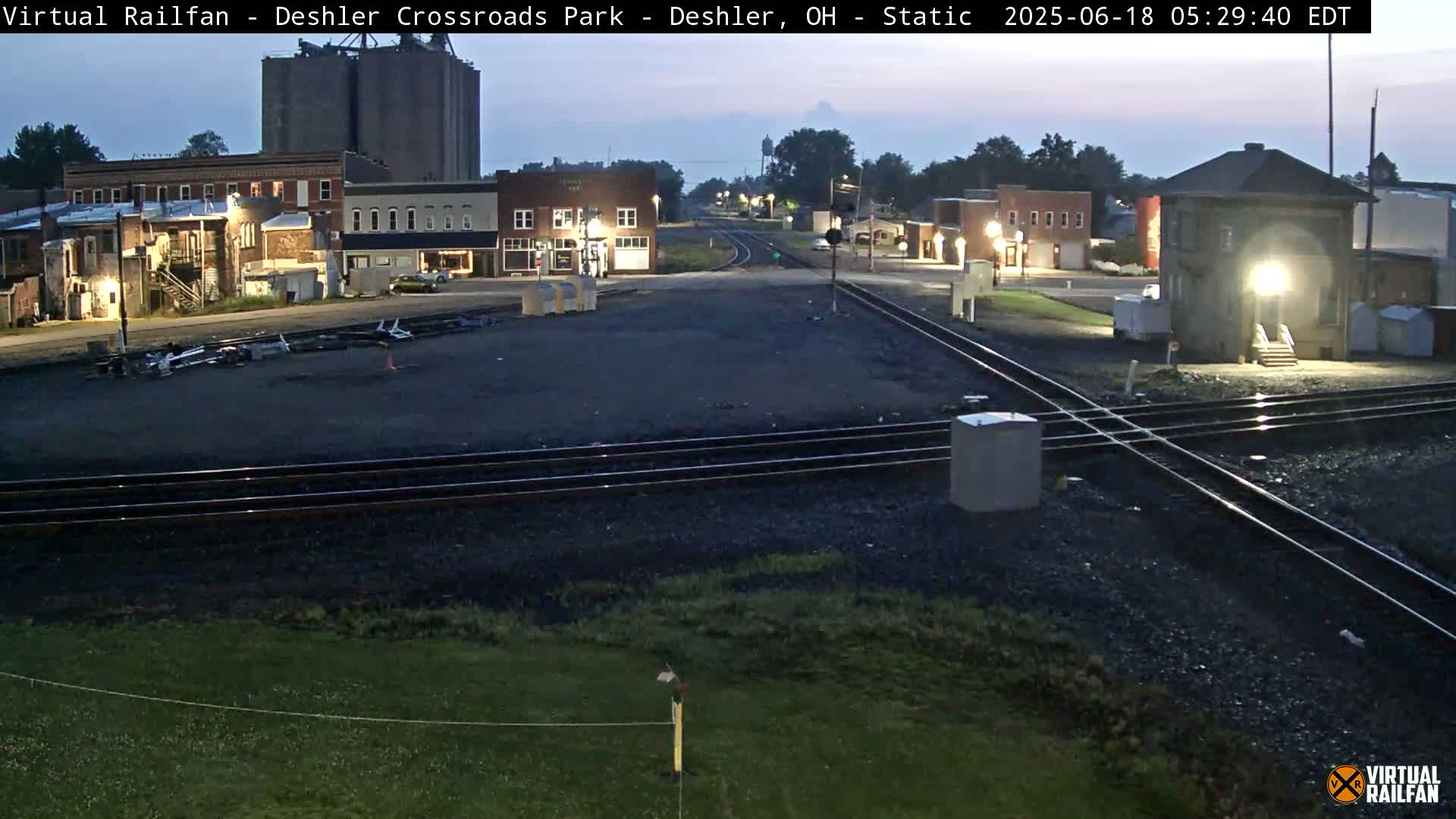A dimly lit, pre-dawn scene shows a railroad crossing in a small town with several buildings, including grain silos, and a control tower.