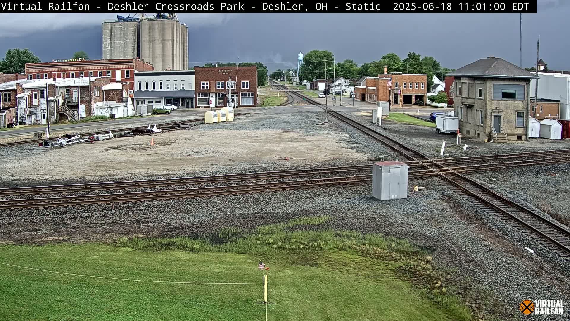 A railroad crossing in a small town under an overcast sky, with several buildings and grain silos visible in the background.