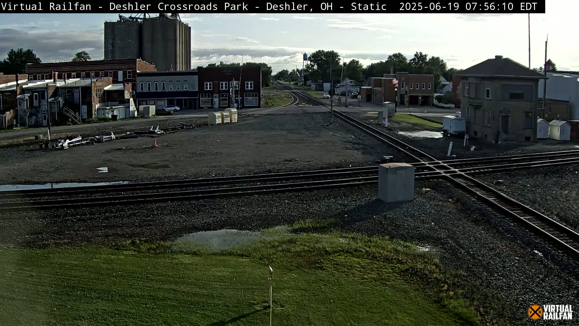 A mostly cloudy day reveals a train crossing in a small town, with several buildings, grain silos, and some puddles visible.