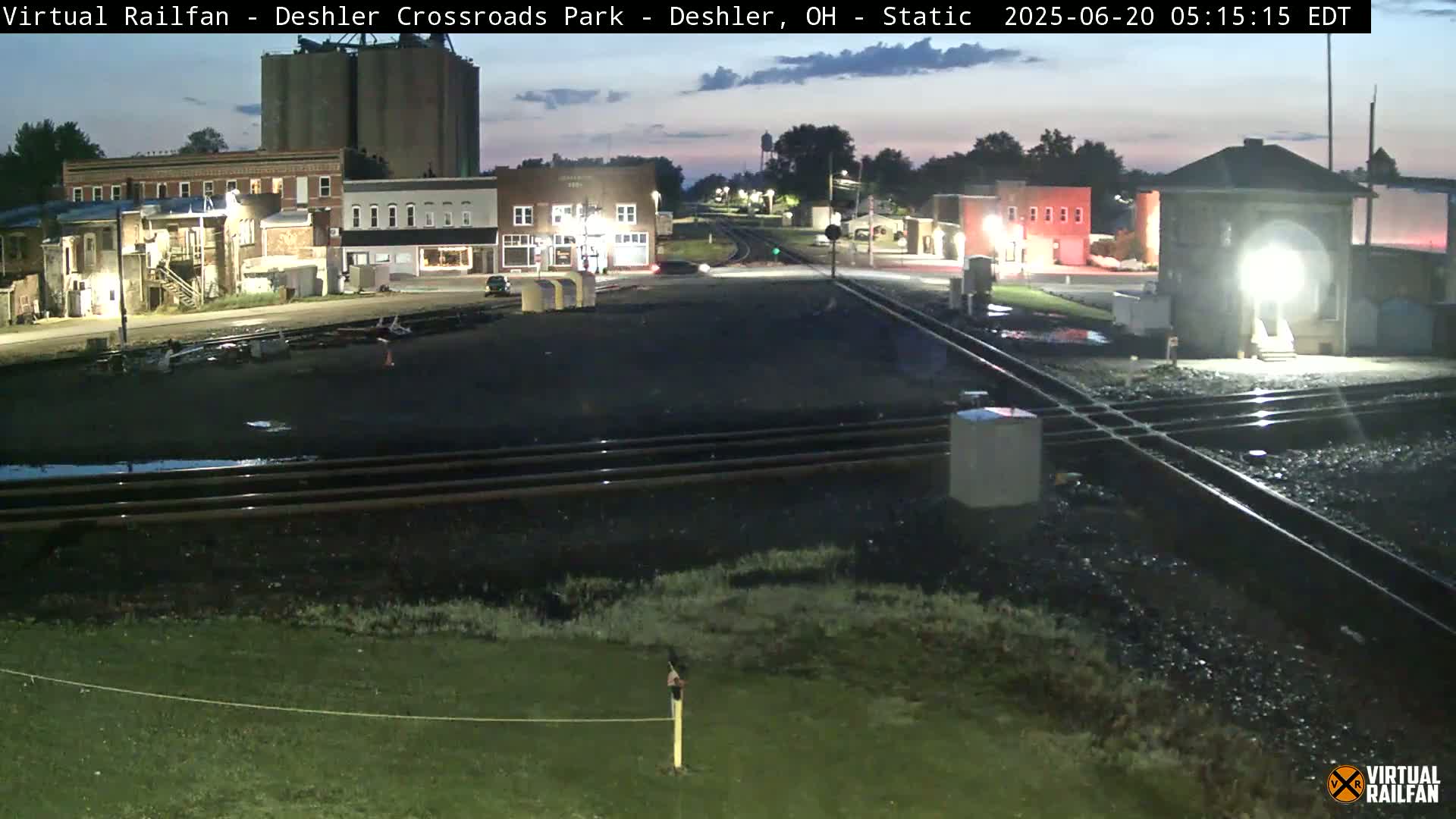 A nighttime view of a railroad crossing in a small town, with several buildings illuminated under a dark, twilight sky.