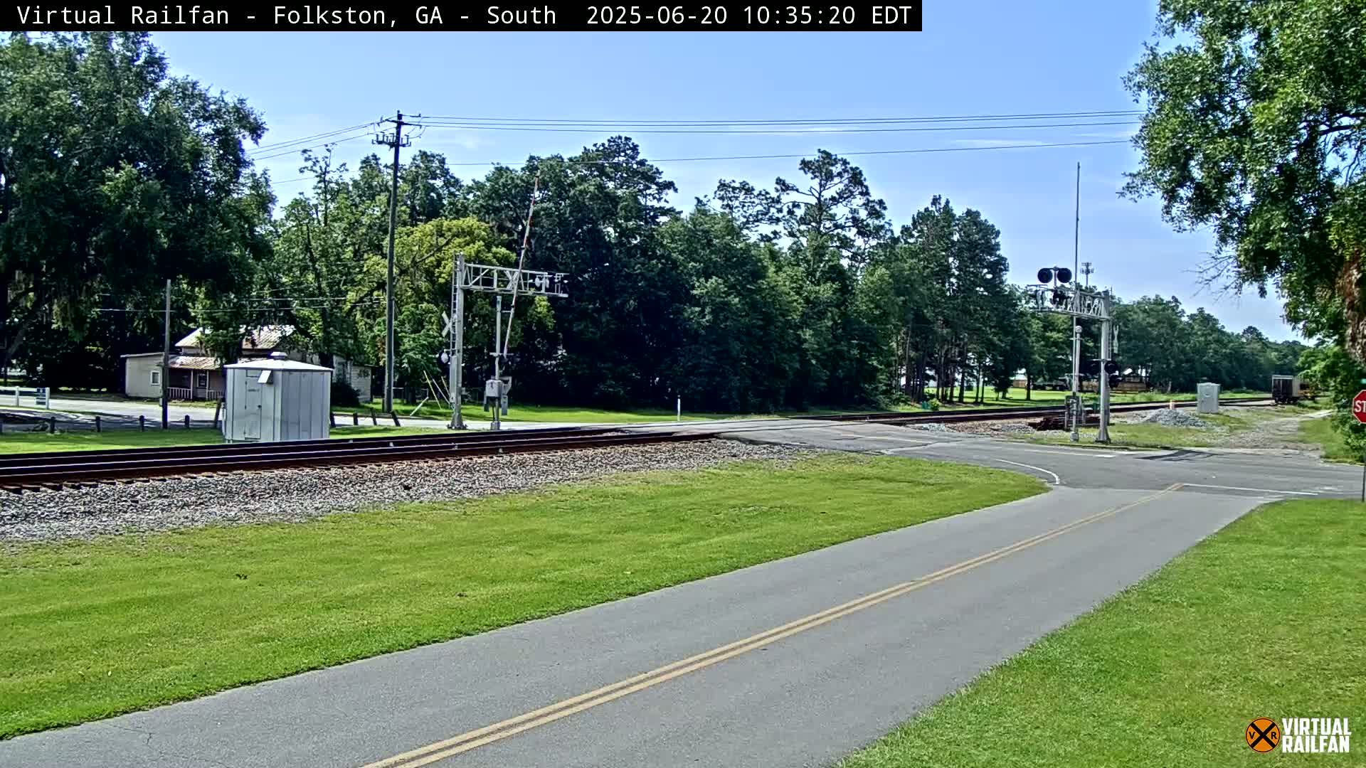A railroad crossing with a road and a paved path intersecting under a mostly sunny sky, surrounded by green grass and trees.