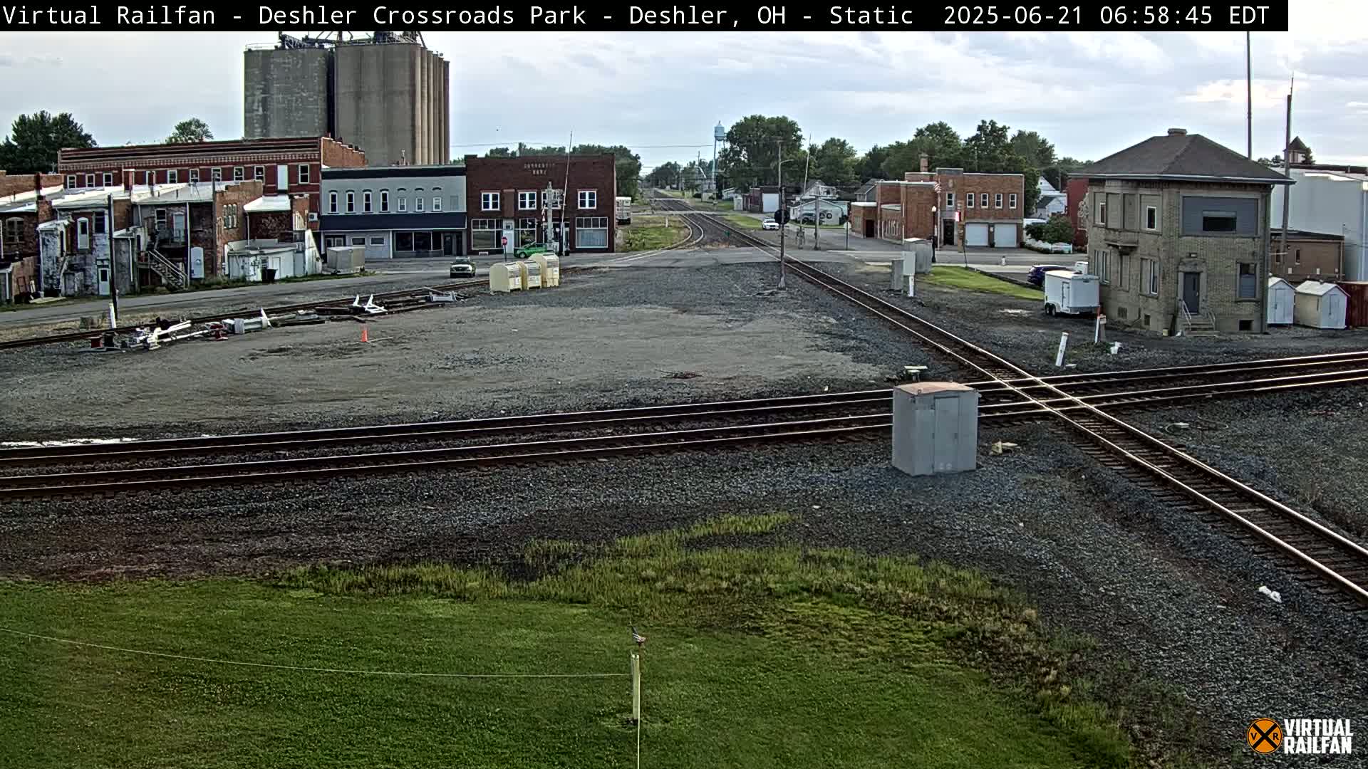 A train crossing in a small town on a partly cloudy day, surrounded by buildings including grain silos and a brick structure, with railroad tracks intersecting in the foreground and grassy areas in the background.