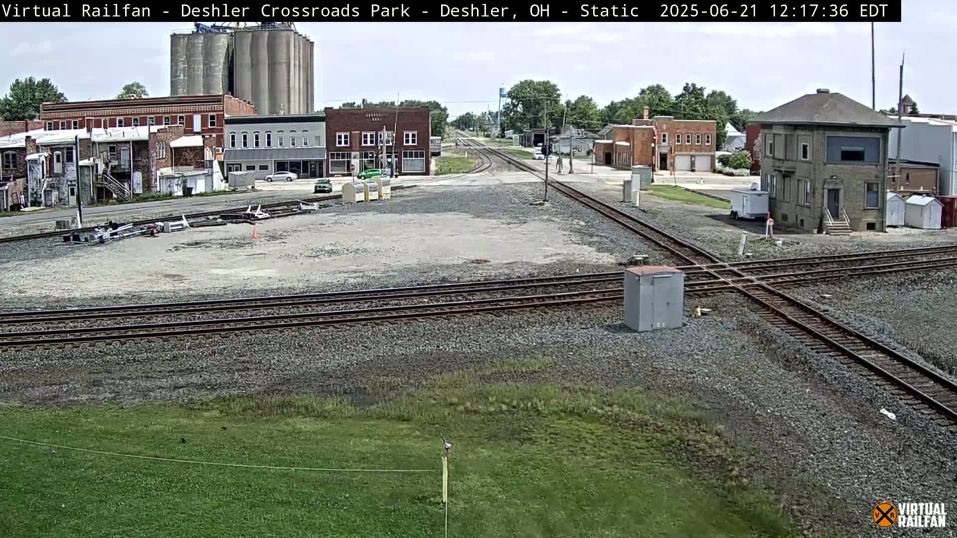 A railroad crossing in a small town on a partly sunny day, with several buildings, grain silos, and railway tracks visible.