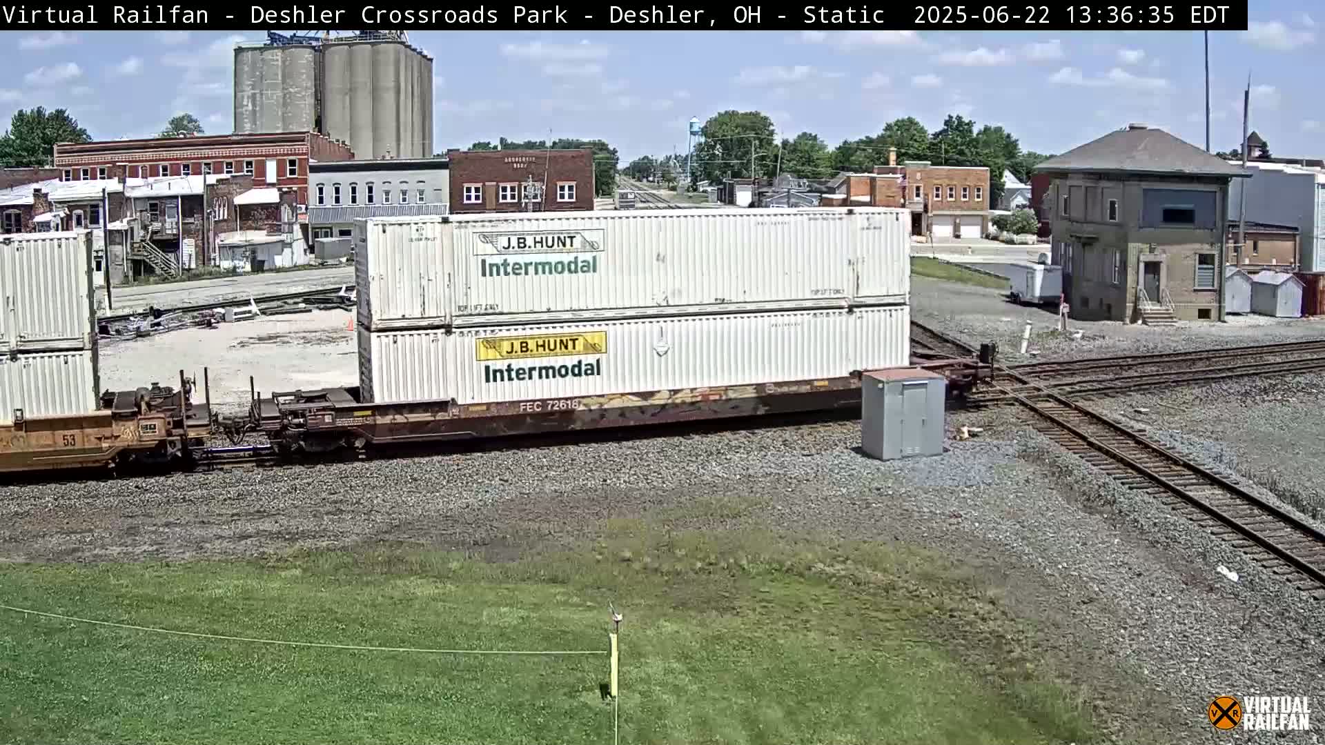 A train carrying two stacked shipping containers sits on a railway track under a sunny sky, with several buildings and grain silos visible in the background.