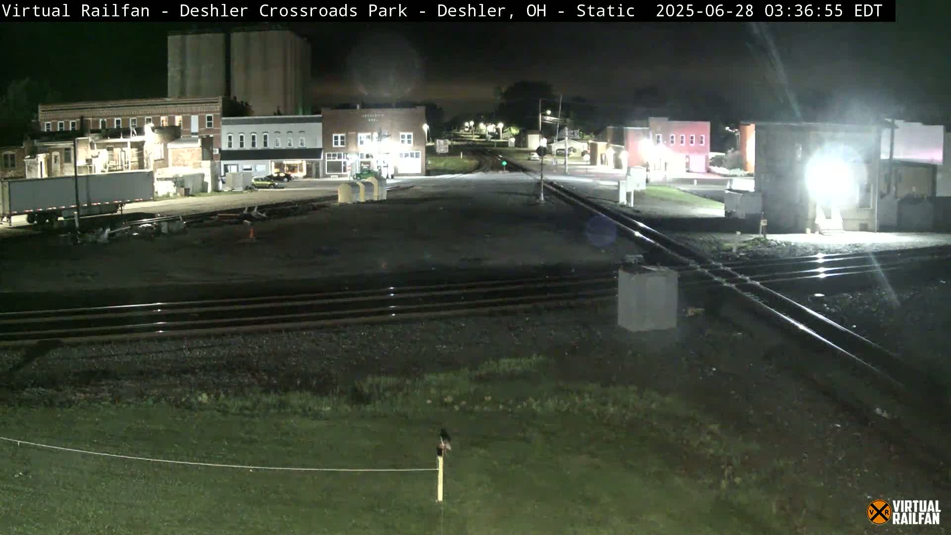 A nighttime view of railroad tracks intersecting a small town, with several buildings and a semi-trailer visible under clear skies.