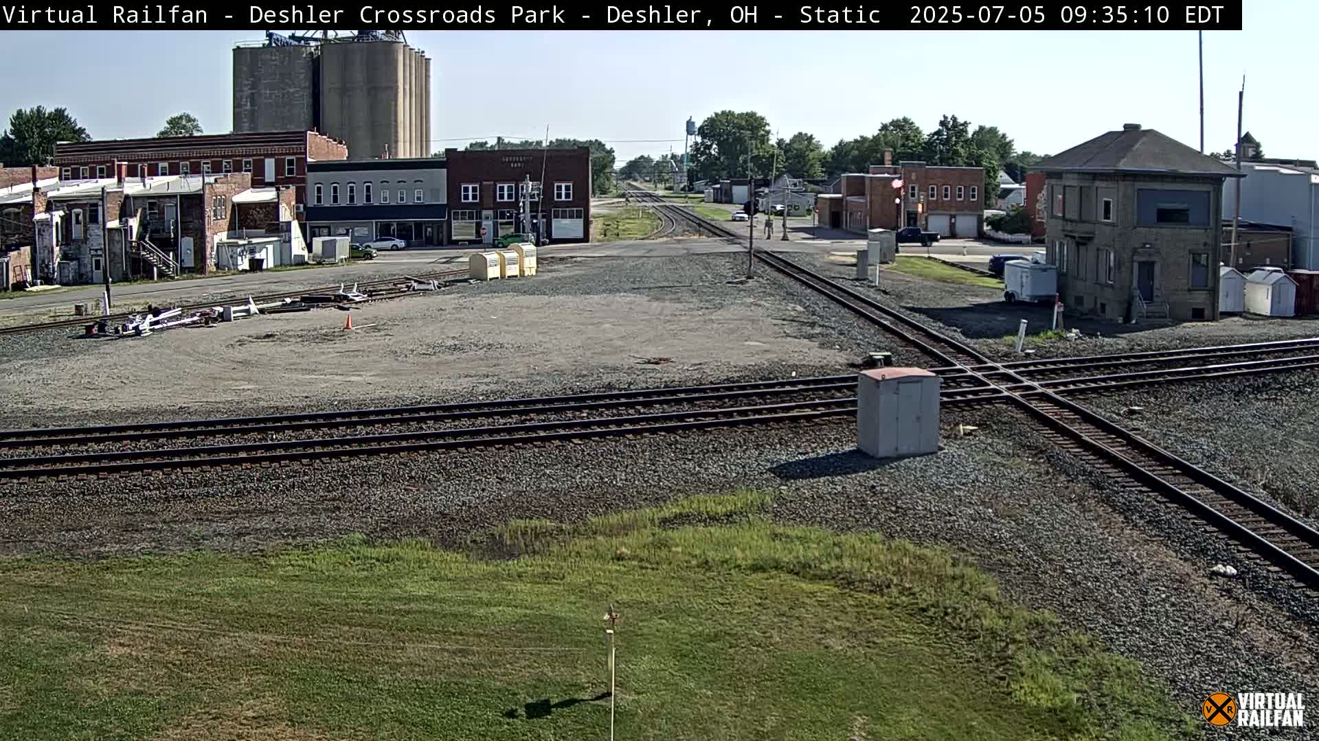 A railroad crossing in a small town on a sunny day, with several buildings, grain silos, and grassy areas visible.