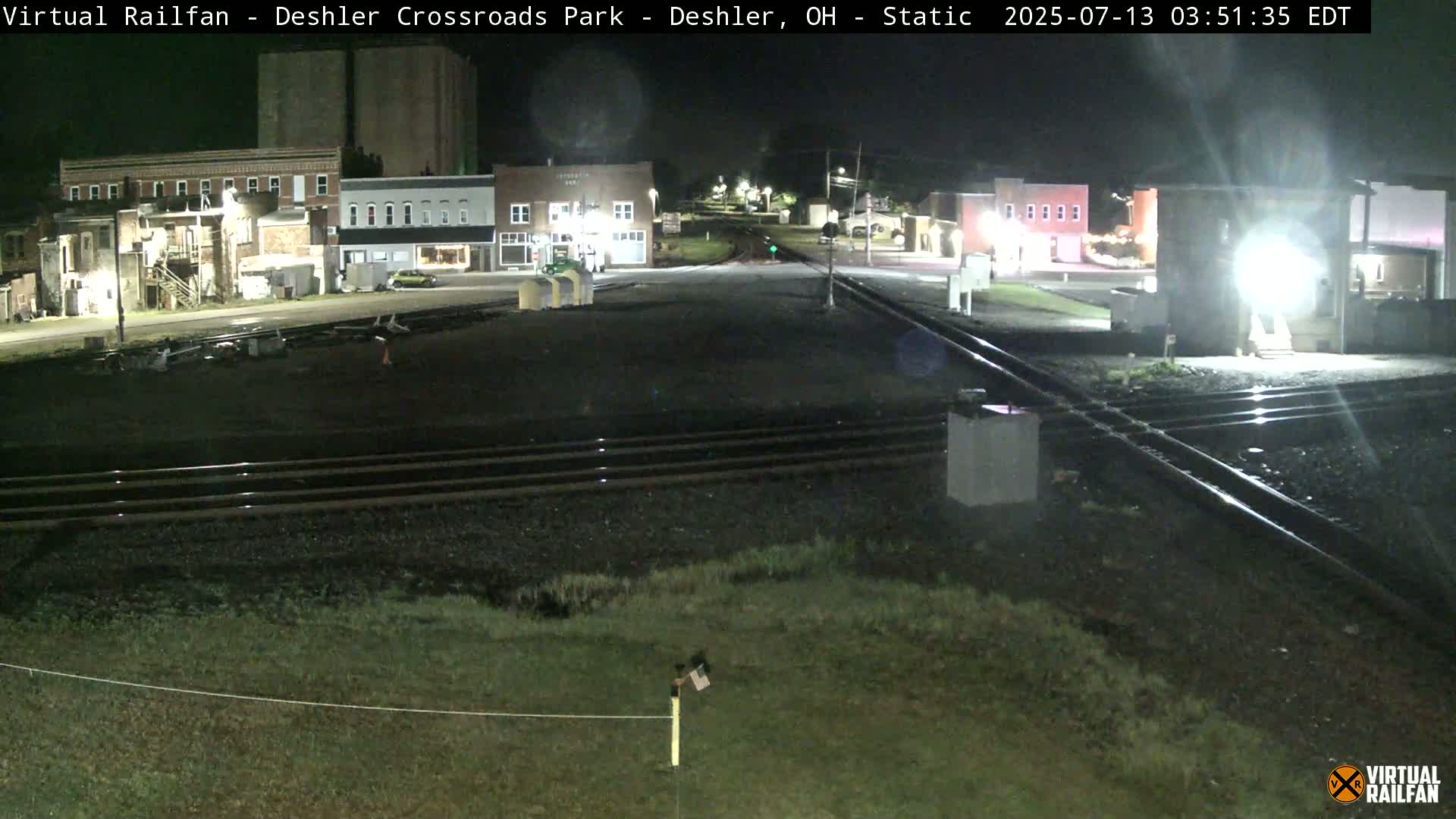 A nighttime view of a railroad crossing in a small town, with several buildings visible under dark, clear skies.