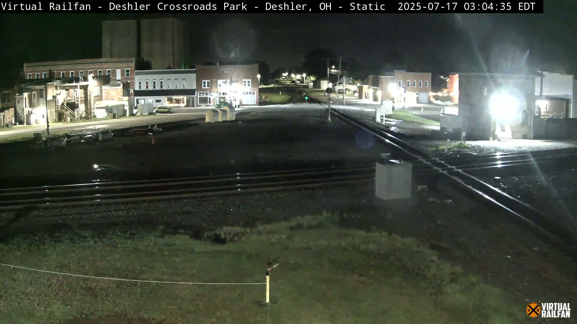 A nighttime view of a railroad crossing in a small town, with several buildings visible in the background.