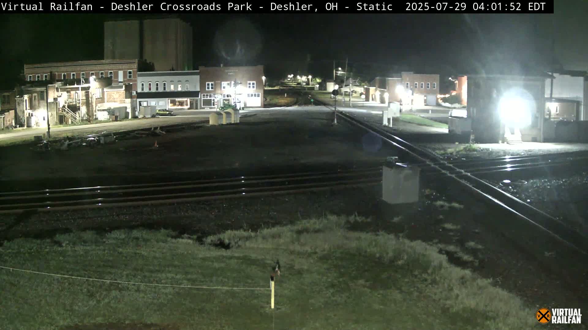 A nighttime view of a railroad crossing in a small town, with several buildings visible in the background.