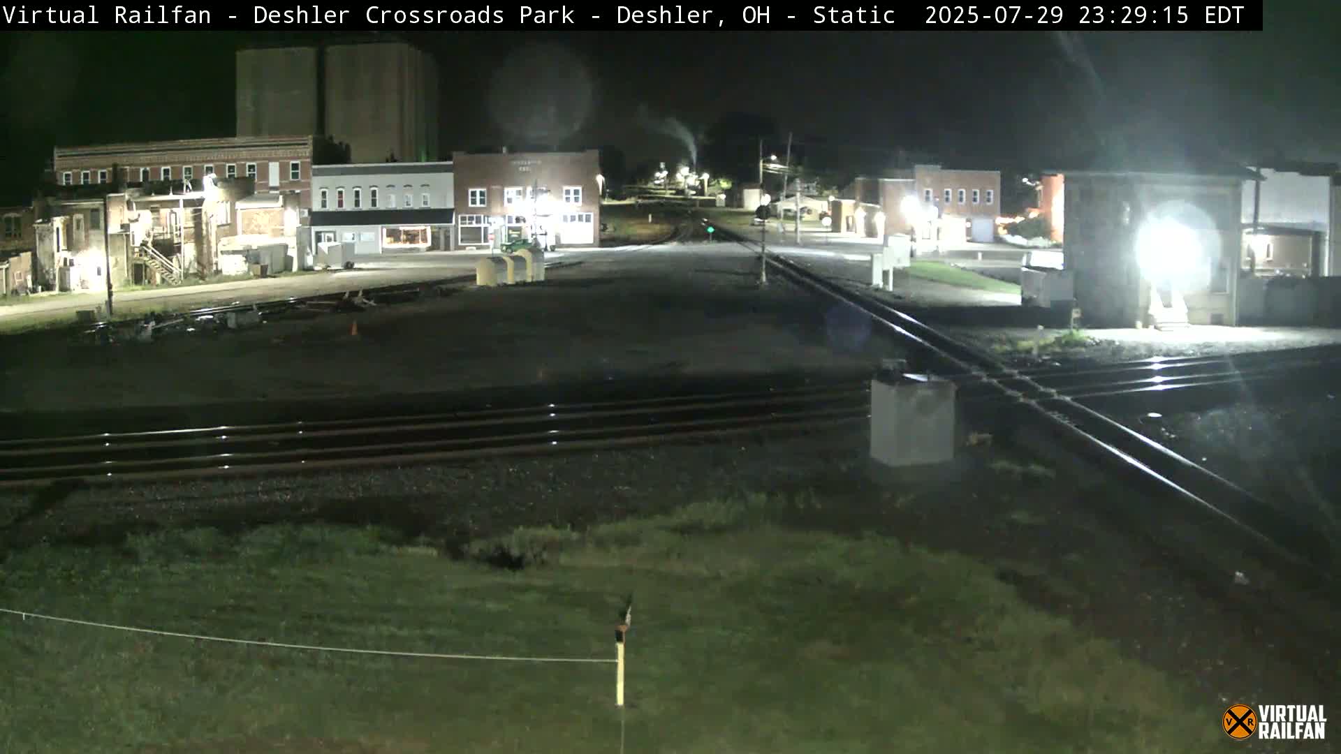 A nighttime view of a railroad crossing in a small town, with several buildings visible in the background.